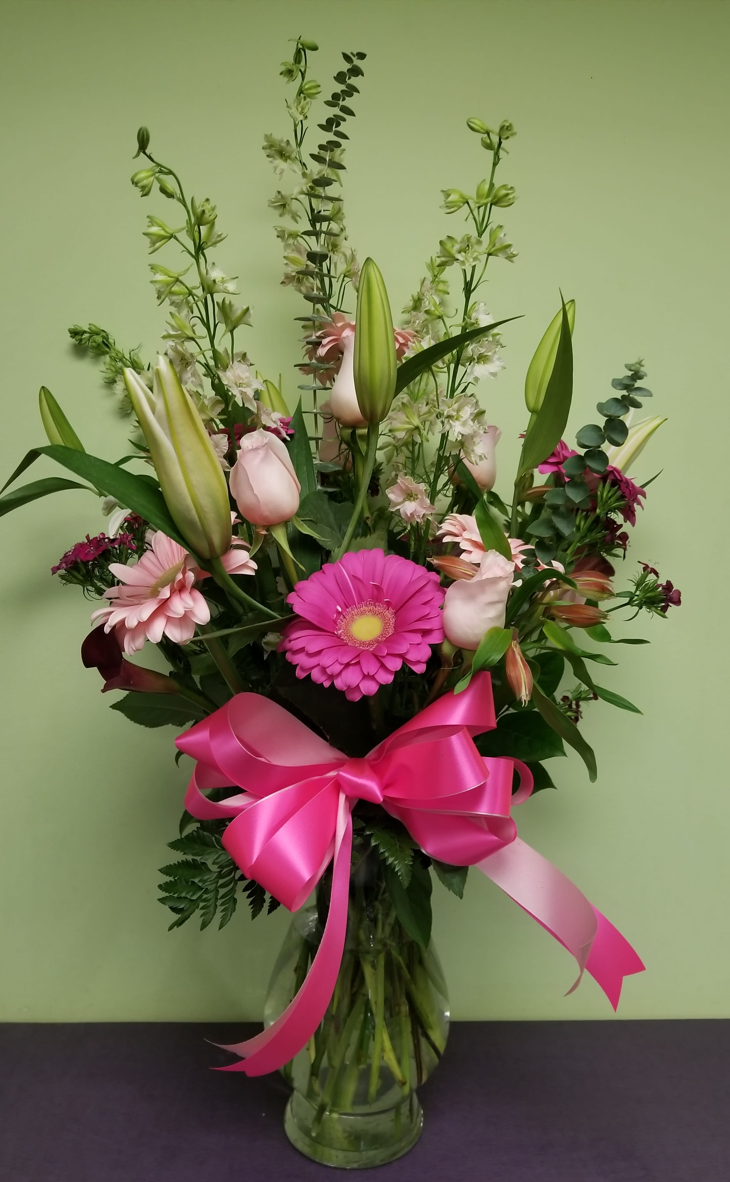 Tall bouquet of pink lilies, gerbera daisies, and a bright ribbon in a glass vase