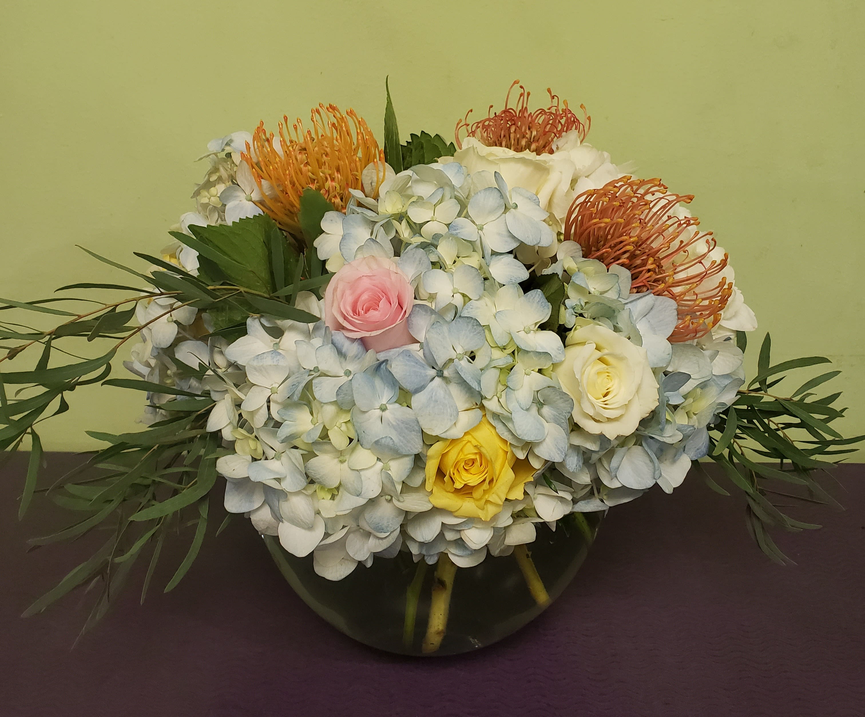 Mixed bouquet of hydrangeas, roses, and orange pincushion blooms in a glass vase