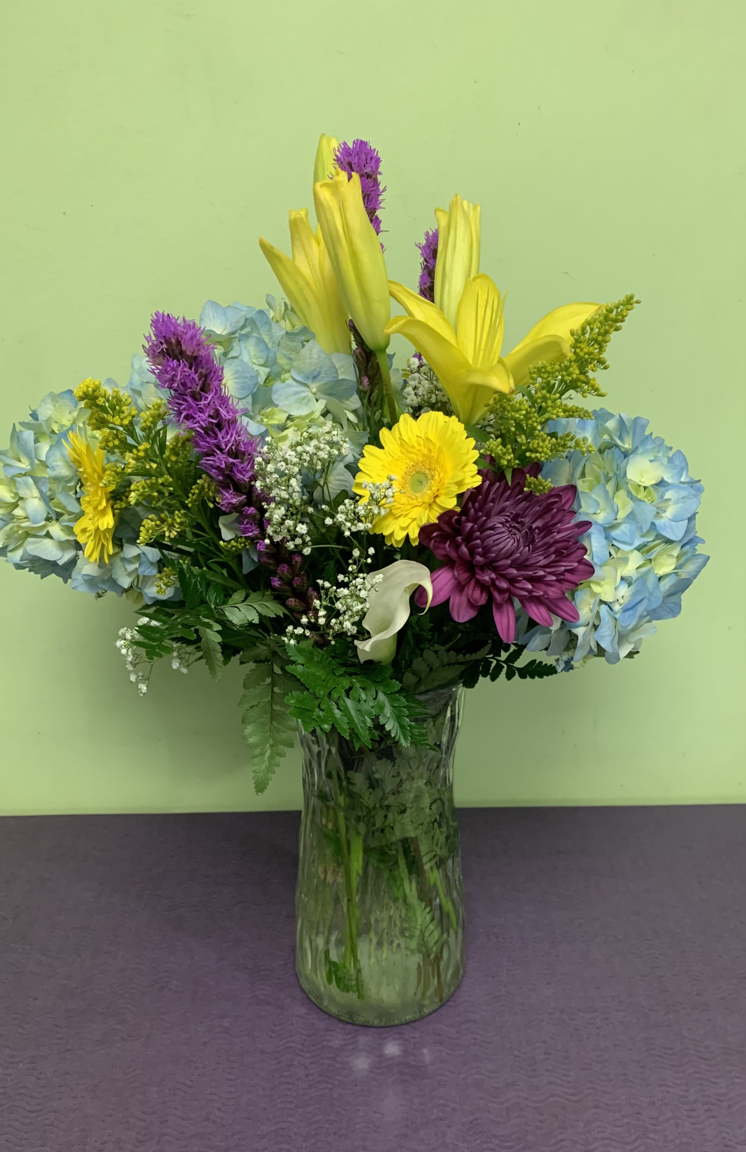 Mixed bouquet in a glass vase with blue hydrangeas, yellow lilies, and purple chrysanthemums