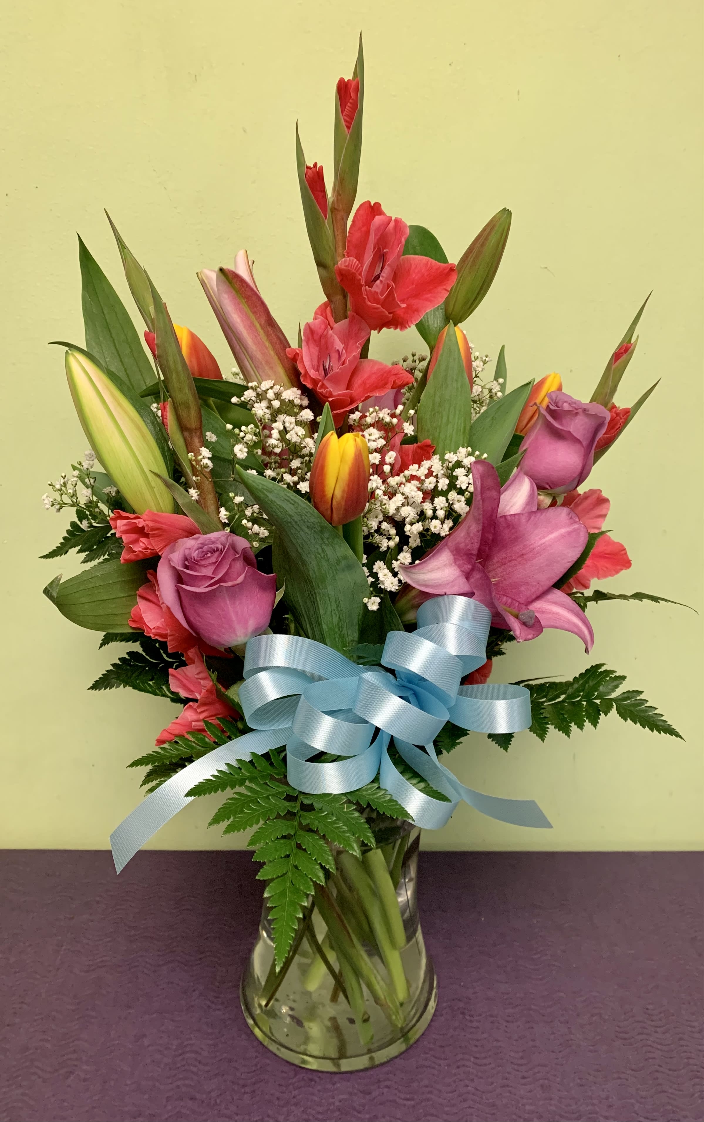 Mixed bouquet of tulips, roses, and lilies in a glass vase with a blue ribbon
