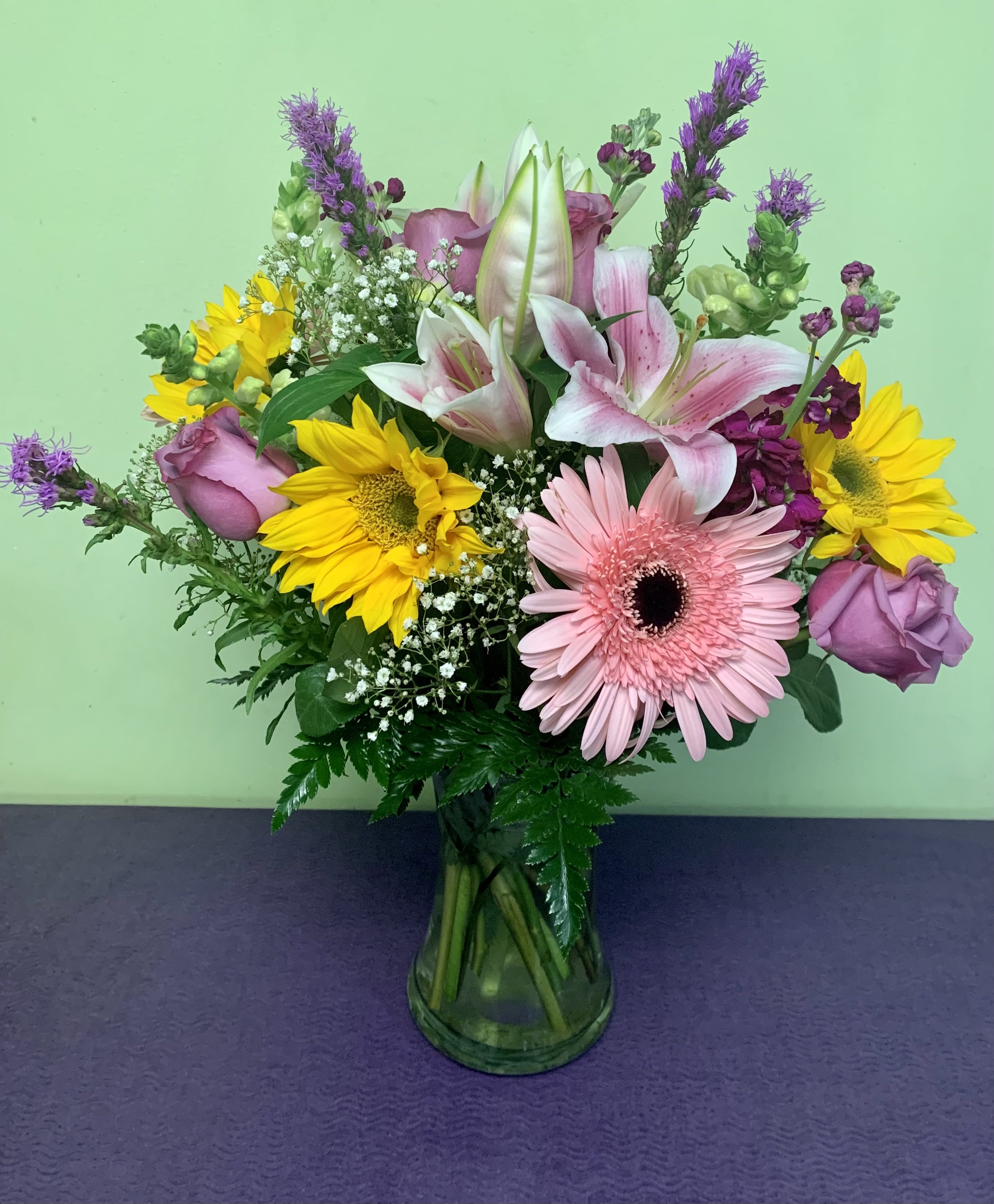 Mixed bouquet of pink lilies, roses, gerbera daisies, and sunflowers in a glass vase