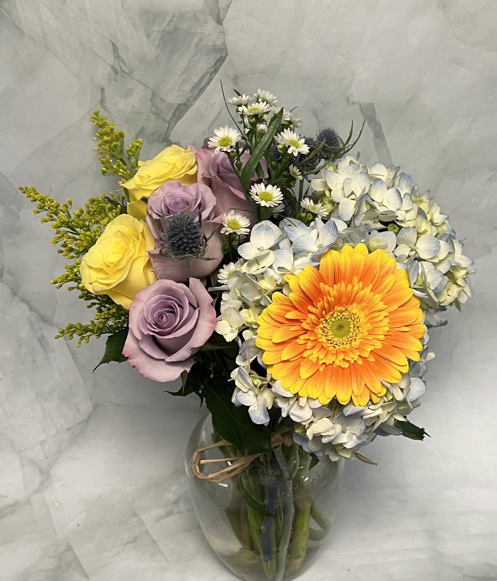 Bouquet of yellow and lavender roses with a bright orange gerbera daisy in a glass vase.