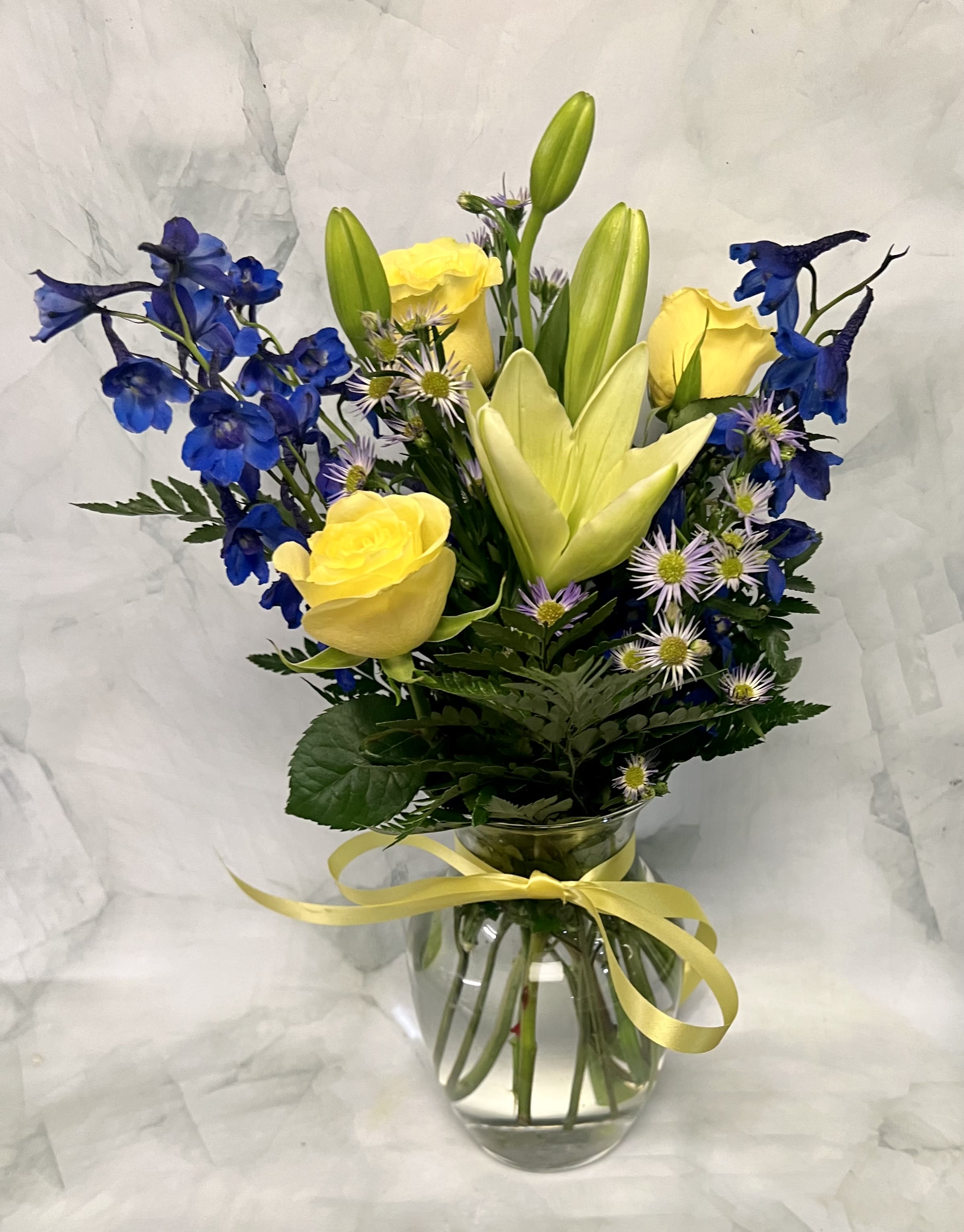Yellow roses and blue delphinium in a glass vase with a ribbon.