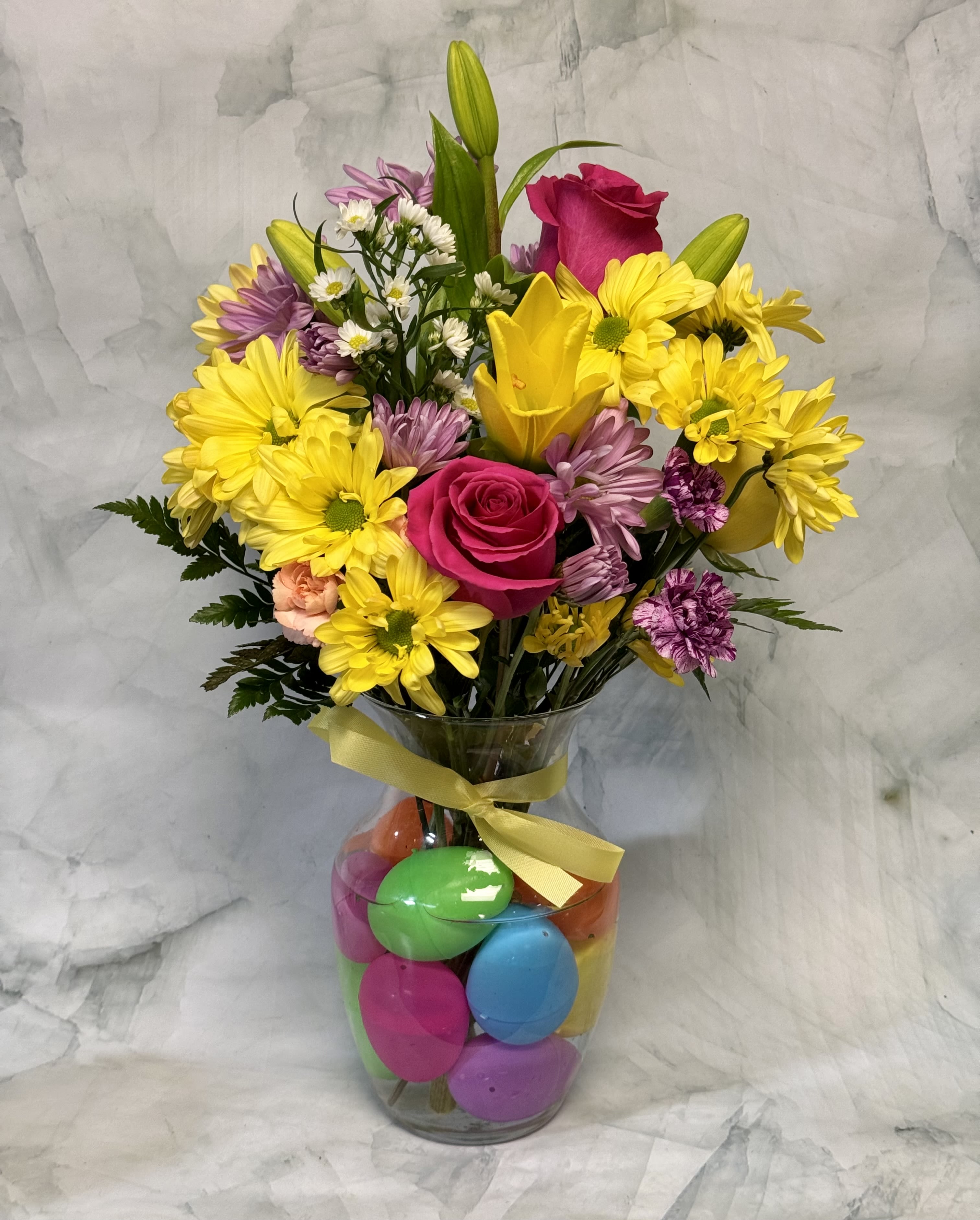 Bouquet of yellow daisies, pink roses, and lilies in a glass vase with colorful eggs