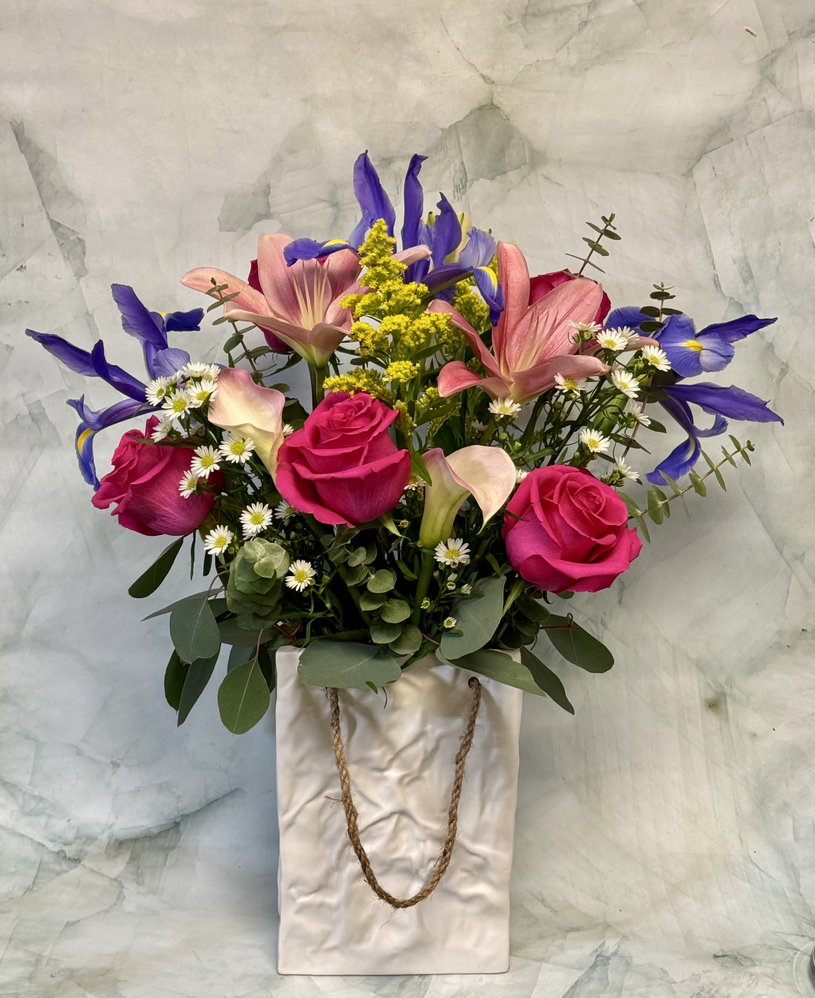 Mixed bouquet of pink roses, lilies, and purple irises in a white ceramic bag-style vase