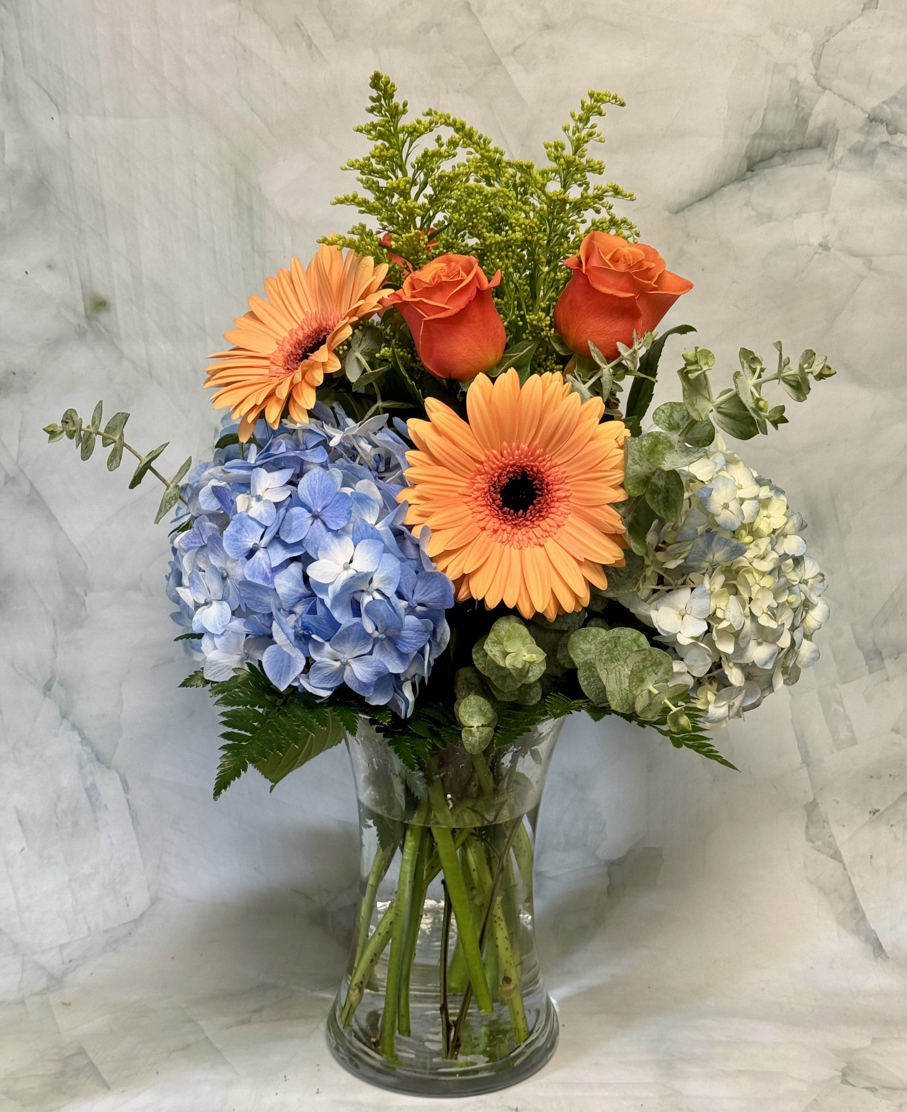 Bouquet of orange gerbera daisies, roses, and blue hydrangeas in a glass vase