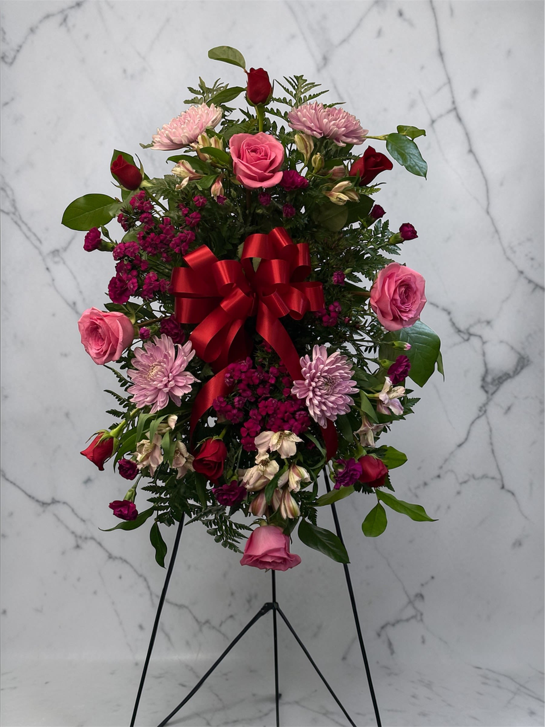 Standing spray of pink and red flowers with a red bow on a black easel.