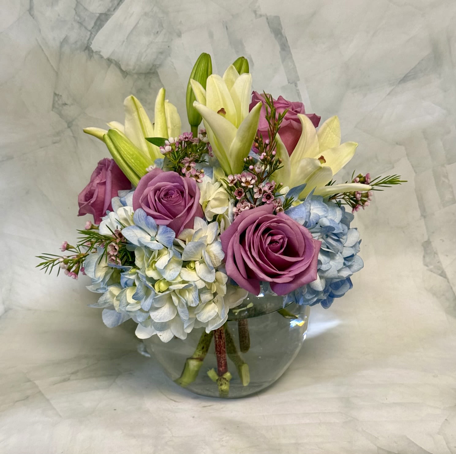 Lavender &amp; Sky Delight - Lavender &amp; Sky Delight greets the recipient with soft, pastel elegance-a mixed-color flower arrangement perfect for a birthday bouquet, anniversary roses alternative, or a cheerful "thinking of you" gift. The combination of lavender roses and pale blue hydrangeas set a calm, romantic tone, while white lilies add a bright, graceful lift so the person you're thinking of will feel cherished and noticed.  This compact arrangement is styled in a low, rounded clear glass vase with blooms packed closely for a full, layered look. Expect silky lavender roses, cloud-like blue hydrangea heads, and creamy white lilies arranged tightly for a polished tabletop presence; the colors read as purple, blue, and soft cream for a fresh, airy mood. Its low shape makes it ideal for dining tables, desks, or bedside surfaces without blocking conversation.  Lovely for celebrations or everyday joy-send it as an anniversary surprise, a birthday pick-me-up, or a considerate way to say you're thinking of someone. The sturdy glass vase and close-packed design help keep stems supported; for best longevity, change the water every 2-3 days and recut stems at an angle when you refresh the display.