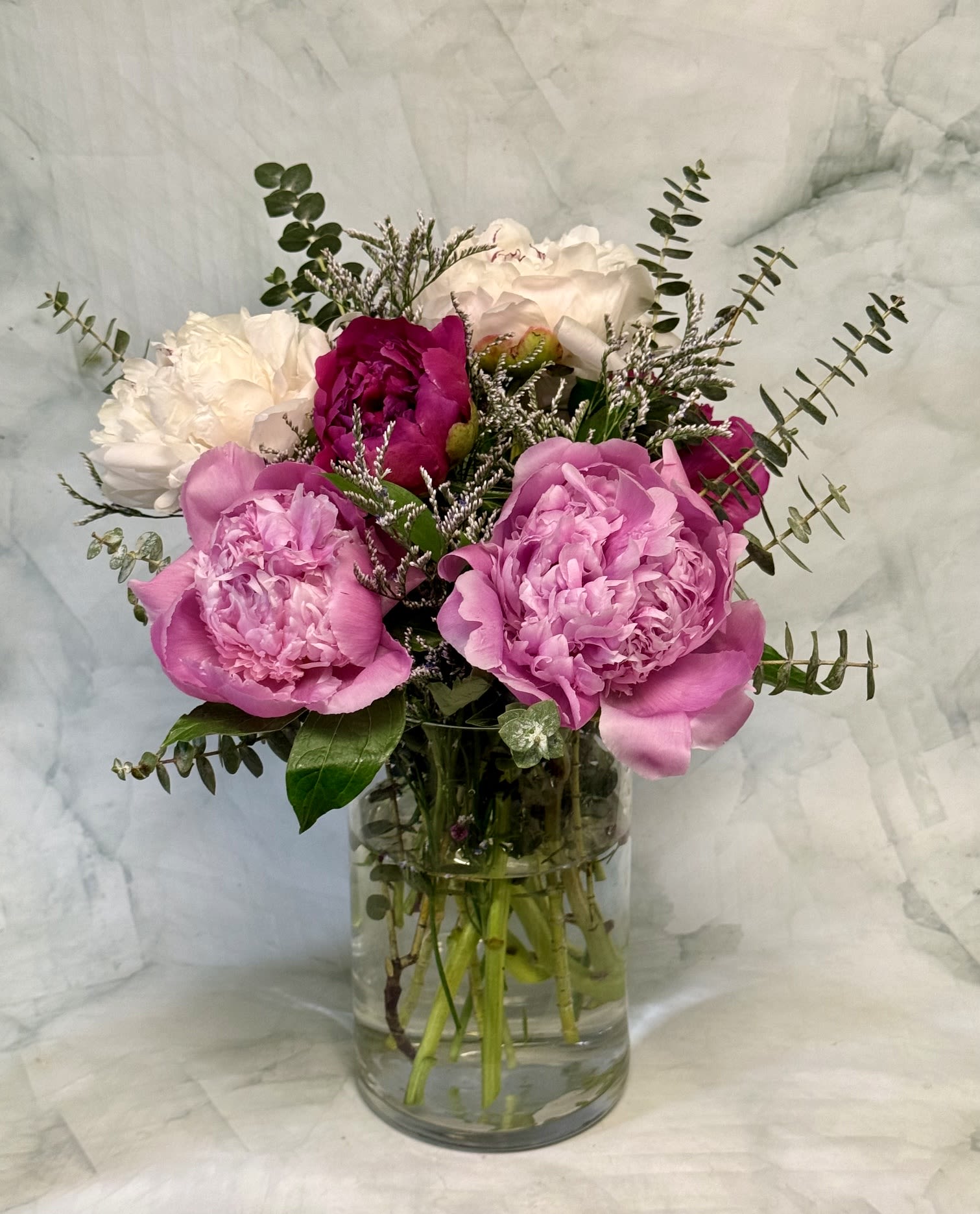Perfect Peonies  - Ruffled pink peonies take center stage in this garden-style flower arrangement, a heartfelt choice for Mother's Day, a birthday, or any moment you want to make Mom feel cherished. The soft pink and white palette reads romantic and fresh, giving the person you're thinking of an immediate sense of calm and delight.  Several large pink peonies are paired with white blooms, fine-textured filler, and airy eucalyptus branches that add height and movement around the fuller flowers. The bouquet is arranged in a clear cylindrical glass vase for a clean, modern presentation that highlights stems and natural texture.  Perfect for the mom who loves a lush, just-picked look, this arrangement makes someone feel truly seen and celebrated-whether you're marking an anniversary or sending an everyday "thinking of you." Delivered ready to display; refresh water regularly and recut stems every few days to keep the peonies opening and lasting longer.  The arrangement includes 6 peonies which may arrive closed. This may cause the arrangement to look smalled that the picture but give the peonies a few days to open. 