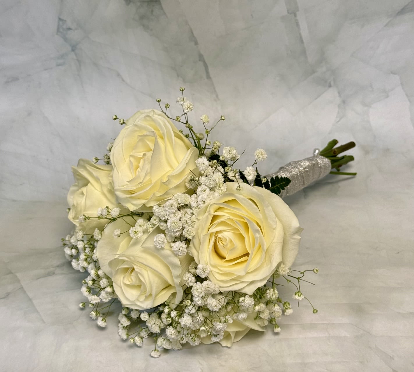 All White Rose Hand Held Bouquet for Prom - White roses gathered into a compact, rounded hand-tied bouquet create an immediate impression of soft romance - perfect as a prom bouquet or a bridesmaid's posy. The arrangement is clearly an all-white/cream rose hand-held bouquet for prom and other special moments where a delicate, understated look is called for; the pale palette reads clean and classic, the kind of bouquet that makes the person receiving it feel cherished and quietly celebrated.  The bouquet centers several cream-to-pale-yellow roses tightly clustered with airy white baby's breath filling the spaces between blooms, giving a gentle, textured contrast of smooth petals and lacy filler. Accents of green foliage lift the composition and keep the palette fresh, while a glitter-wrapped handle adds a subtle, formal finish that's easy to carry. The small, intimate scale and rounded silhouette make it ideal for close-up moments and photos without overwhelming the wearer.  Hand-tied for a neat, durable finish and finished with a decorative glitter handle, it's ready to hold and enjoy right away; treat it as a delicate bloom set and trim stems slightly and keep in fresh water to extend vase life.