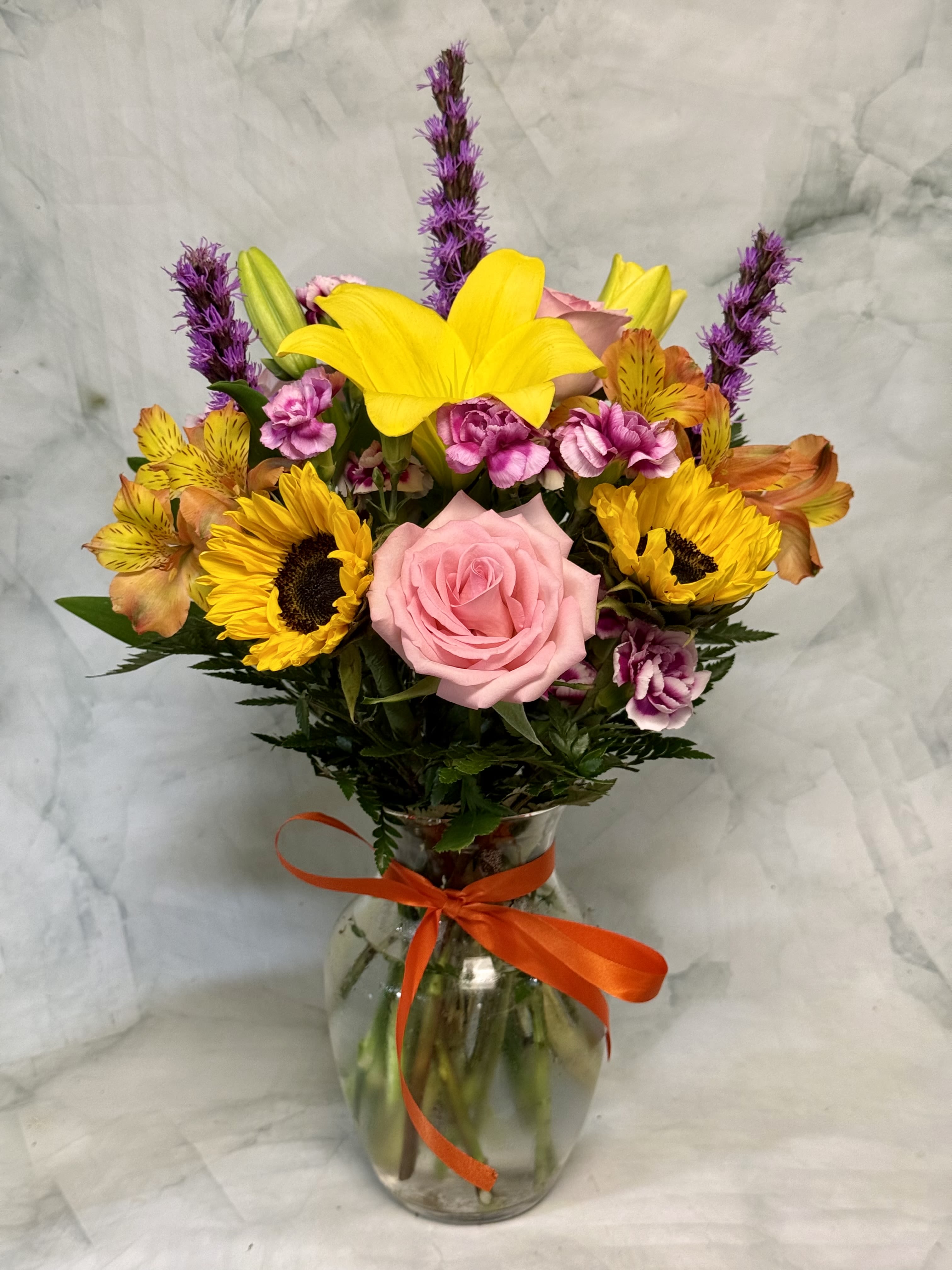 A Summer Sunset - Bright, warm sunflowers paired with a single pink rose create a striking focal point in this summer bouquet - perfect as a graduation gift, a heartfelt "I'm sorry," or a bright "thank you" to someone who needs lifting. A Summer Sunset is a flower arrangement that reads like a sunset in bloom: warm oranges and yellows balanced by cool purples for contrast, giving the recipient an immediate sense of cheer and calm.  This arrangement features yellow sunflowers, a central pink rose, small pink-and-white carnations, orange alstroemeria, purple accent spikes (lending height and texture), daisies, asiatic lilies, and complementary dianthus gathered in a clear glass vase tied with an orange ribbon. The hand-tied composition sits neatly in the vase for easy display; colors range from golden sunflower yellow through coral and orange to lavender purple, creating layered visual depth and a lively, upbeat silhouette.  Give this bouquet to someone celebrating summer milestones, graduation, or to say thank you or I'm sorry - it's the kind of arrangement that makes the recipient feel noticed and celebrated. Vase-arranged for immediate enjoyment; trim stems and refresh water every 2-3 days to extend bloom life.