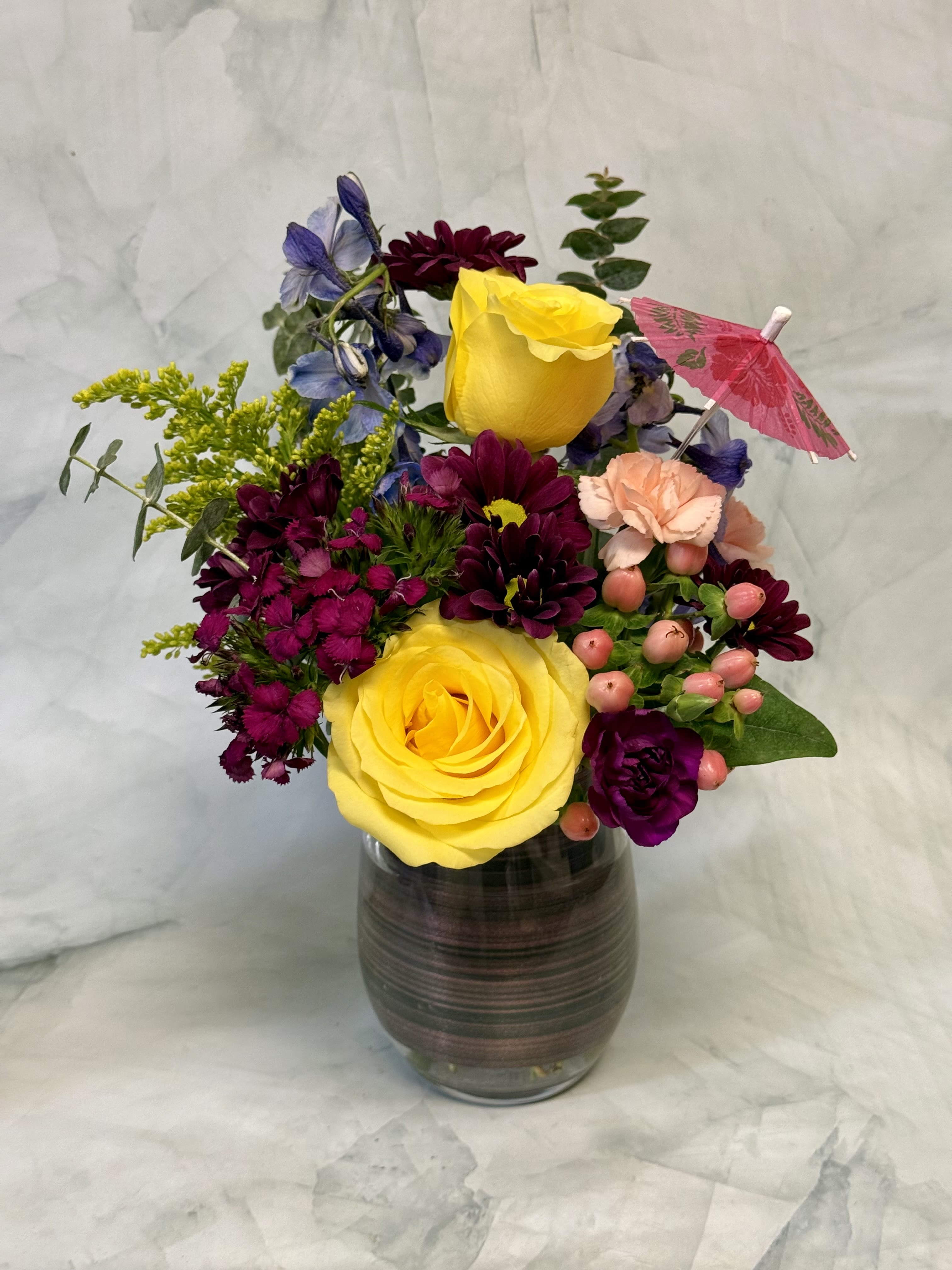 Happy Hour Flowers - Raise a glass to life’s special moments with Happy Hour Flowers, a playful and vibrant floral arrangement designed in a charming wine glass and topped with a festive cocktail umbrella. This unique design brings together the fun of happy hour and the beauty of fresh flowers, creating a gift that’s sure to make anyone smile.  Featuring sunny yellow roses, rich burgundy daisy mums, delicate blue delphinium, soft peach carnations, pink hypericum berries, and textured seasonal accents, this arrangement is bursting with color and personality. The bright yellow roses symbolize friendship, joy, and happiness, while the jewel-toned blooms add depth and a touch of sophistication. Nestled in a keepsake wine glass, the flowers create a compact yet eye-catching display that’s perfect for a desk, kitchen counter, dining table, or bar area.  Whether you’re celebrating a birthday, retirement, promotion, girls’ night, summer gathering, or simply sending a cheerful surprise, Happy Hour Flowers delivers a refreshing splash of color and fun. The whimsical cocktail umbrella adds a tropical-inspired finishing touch, making this arrangement feel like a mini vacation in a glass. It’s the perfect way to say “cheers” and brighten someone’s day with fresh flowers and festive flair.