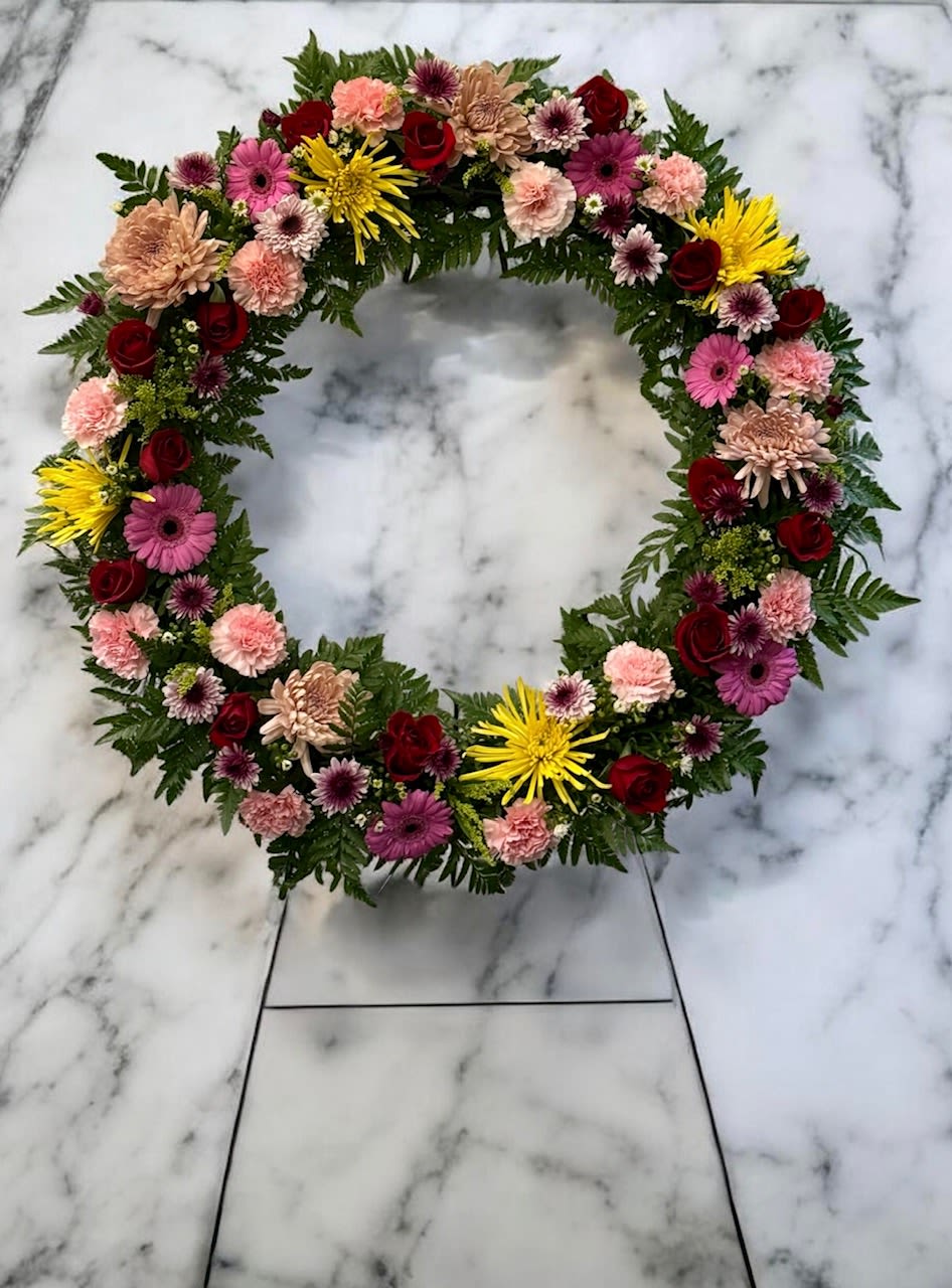 Colorful Sympathy Wreath - A richly layered, circular sympathy wreath designed to convey warmth and remembrance-this Colorful Sympathy Wreath is a formal memorial arrangement ideal for a funeral service or graveside tribute. The mixed-color palette of pinks, reds, yellows, whites, and peaches reads as both comforting and celebratory, making it a thoughtful choice when you want the person you're remembering to be honored with color and care.  Dense clusters of roses, carnations, gerbera daisies, and chrysanthemums cover the wreath frame for a full, textured surface; dark green foliage around and beneath the blooms provides a strong base that lets the brighter flowers pop. The arrangement is built on a traditional wreath frame for a balanced, symmetrical silhouette that lays flat or stands on an easel-its formal, commemorative shape communicates respect and lasting sentiment.  Choose this wreath to express sympathy, celebrate a life at a memorial service, or leave at a gravesite-she'll feel genuinely seen by the warmth of the colors and the thoughtful construction. Because it's assembled on a wreath frame, it's ready for display upon delivery; simple care like keeping it sheltered from harsh sun and wind will help preserve the blooms for the service.