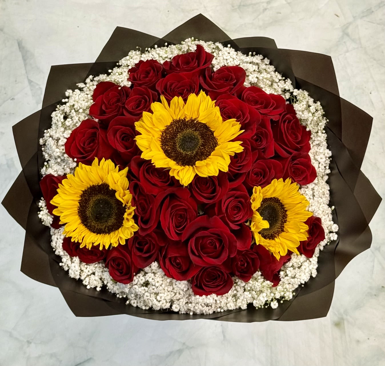 The Ramos - A dense cluster of deep red roses takes center stage in this romantic flower arrangement, with three bright yellow sunflowers cutting across the front to add warmth and cheer - perfect as a birthday bouquet, a love and romance gift, or an everyday surprise for someone you adore. The contrast between velvety crimson roses and sunny sunflower faces creates an immediate emotional lift: she'll feel both cherished and genuinely celebrated the moment she sees it.  Small white filler blooms frame the roses and sunflowers, softening edges and adding a delicate, airy texture around the rounded shape. The stems are finished in dark brown wrapping paper that gives the design a modern, grounded feel and makes the colors pop. Sized to read as full and generous, this arrangement is ready to present without extra fuss and suits recipients who love classic roses with an unexpected, joyful twist.  Includes 30 red roses, 3 sunflowers and babies breath. 
