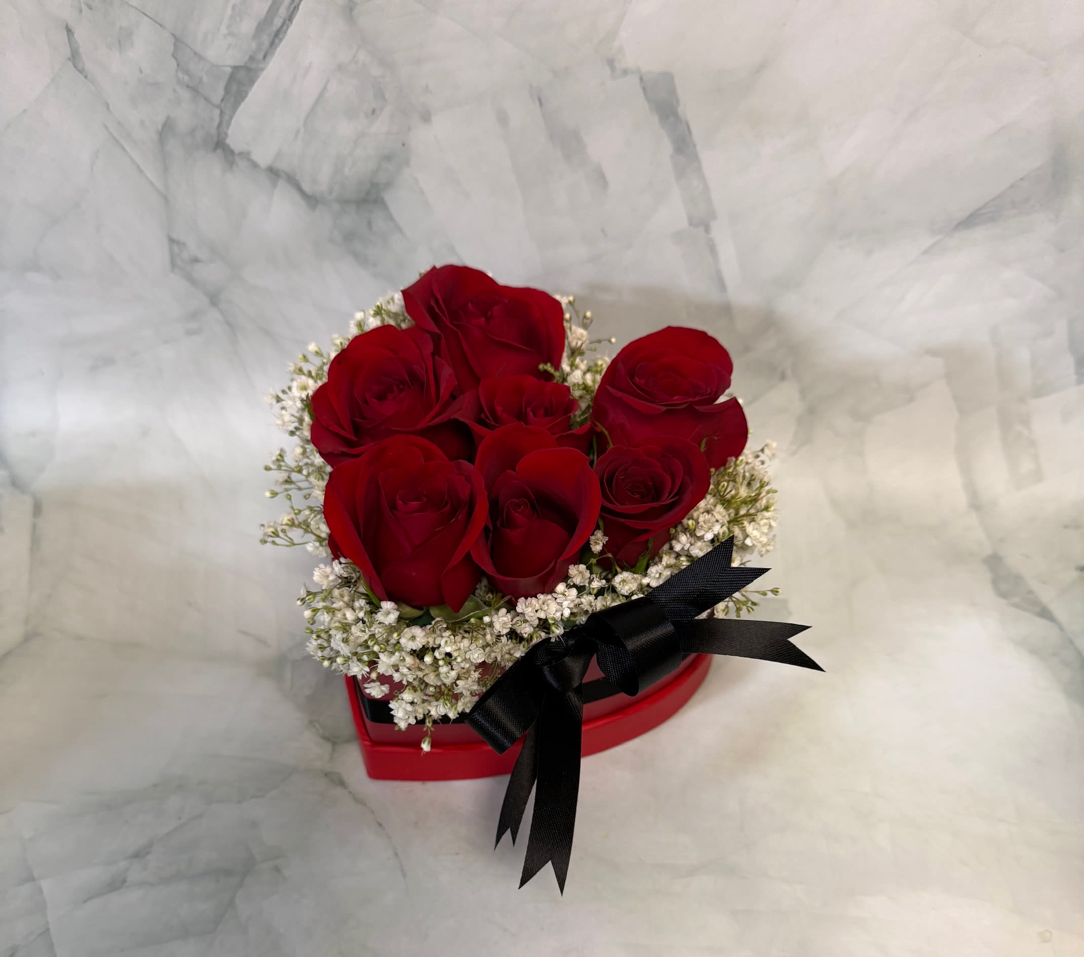 Red roses arranged in a heart-shaped box with white filler flowers and a black ribbon