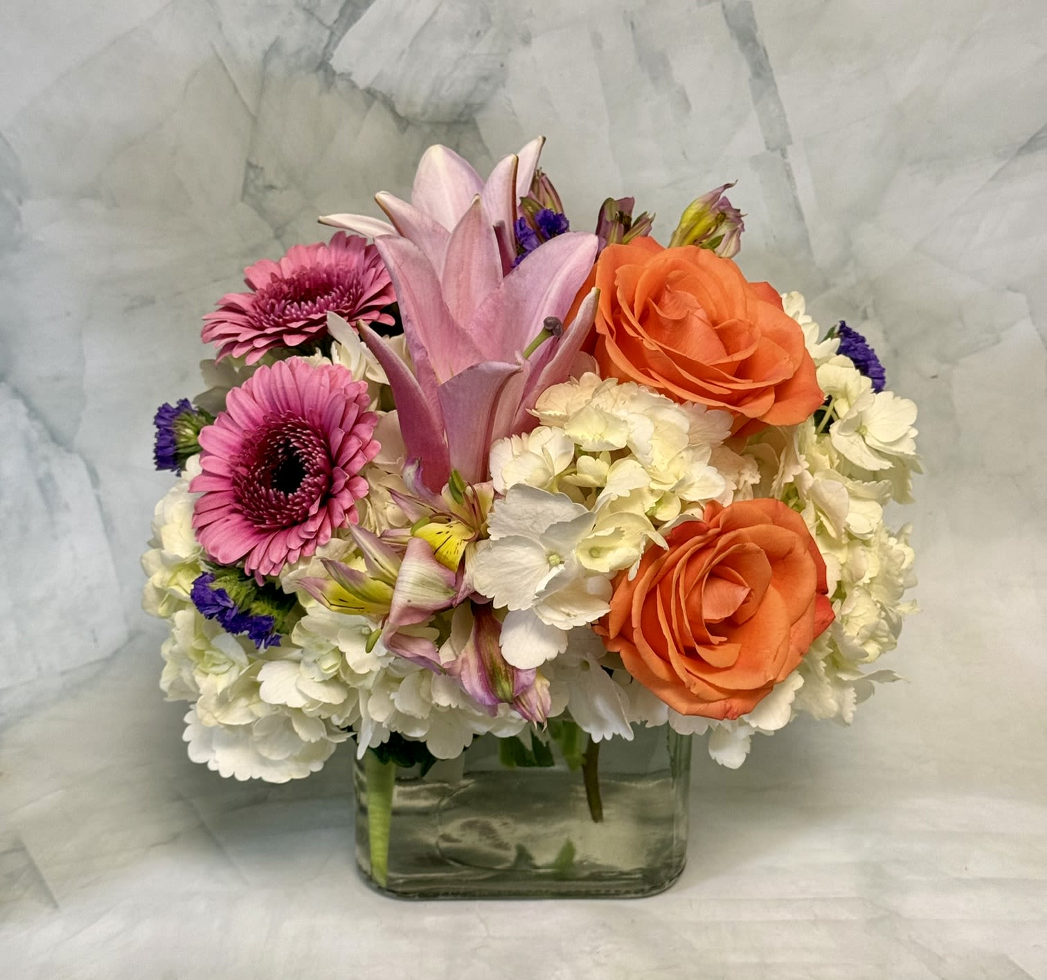 The Beauty of Spring - A bright mix of warm orange roses, pink gerbera daisies and pale pink lilies set against clouds of white hydrangea makes this birthday or "I'm sorry" bouquet feel both cheerful and genuinely heartfelt. This spring-ready flower arrangement and wedding table accent arrives as a low, rounded design in a clear square glass vase so the recipient sees stems and blooms from every angle - the orange and peach tones bring warmth while pinks, purples and whites keep the look soft and romantic.  Packed tightly for a full, balanced presentation, the arrangement also includes alstroemeria, asters and small purple accent blooms that add texture and delicate contrast. Dimensions and vase style create an easy centerpiece for Easter or Passover gatherings, a thoughtful congrats gift, or a polished pick for Administrative Professionals' Week. To keep the bouquet fresh, place in bright, indirect light and top up water daily; the clear glass vase makes refreshing simple.