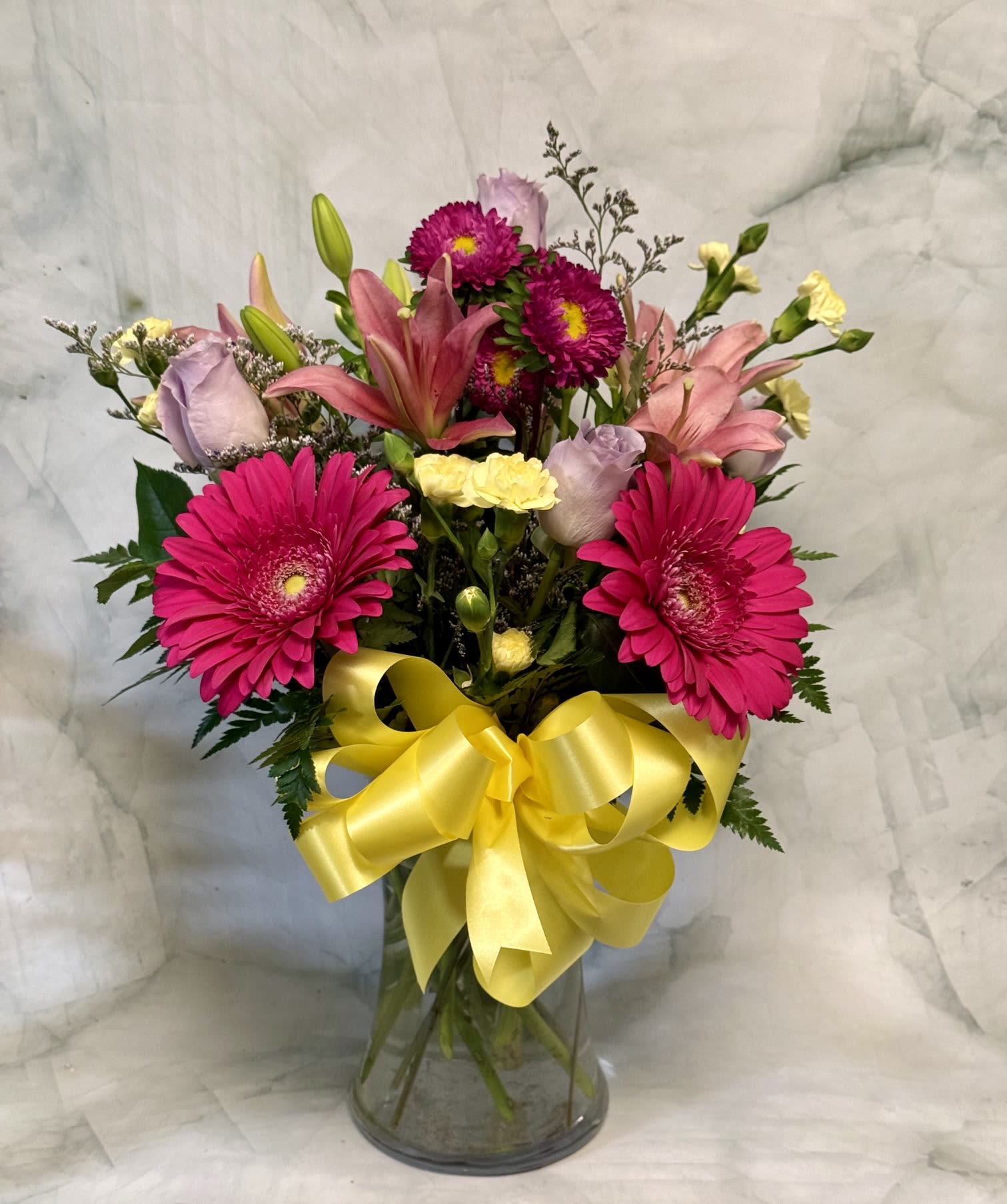 Vibrant Floral Spring Bouquet - Bright magenta and pink gerbera daisies take center stage in this spring flower arrangement, paired with orange Asiatic lilies and soft lavender roses to create a joyful birthday bouquet or a cheerful Admin Professionals' Week gift. The mix of saturated and pastel tones-pale peach and yellow carnations, purple lisianthus and asters-reads lively and celebratory, the kind of bouquet that makes the person you're thinking of feel truly seen.  Arranged in a clear glass vase and finished with a large yellow satin ribbon, the design is full and rounded with broad green leaves framing the blooms and fine filler flowers adding airy texture. Ideal for brightening a desk, entryway, or bedside table; available in a single upright vase size that ships gift-ready with simple care instructions to keep blooms fresh.