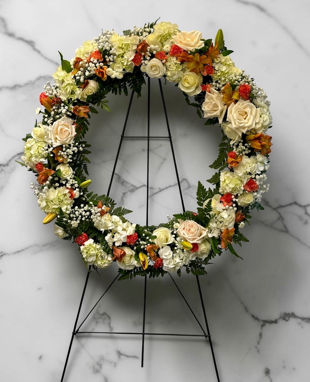 Orange and White Standing Wreath - Cream and peach roses and hydrangeas form a warm, autumn-leaning circle in this standing wreath-perfect as a sympathy wreath or celebration-of-life tribute. The arrangement's orange-red accents and soft white filler flowers create a comforting, layered texture that reads as both formal and welcoming for memorial services or ceremonial displays.  This circular floral wreath is built full and balanced on a sturdy black metal easel stand, sized to read clearly at the front of a service or event. The palette blends white, cream, soft peach, orange and hints of pink and yellow, with roses, hydrangeas, carnations, daisies and mixed filler blooms arranged for depth and a natural, textured silhouette.  Made to honor and console, this standing wreath sends a thoughtful, respectful message-an elegant way to mark a life or a season. Place orders with event timing in mind so we can ensure fresh blooms and proper delivery to the venue.