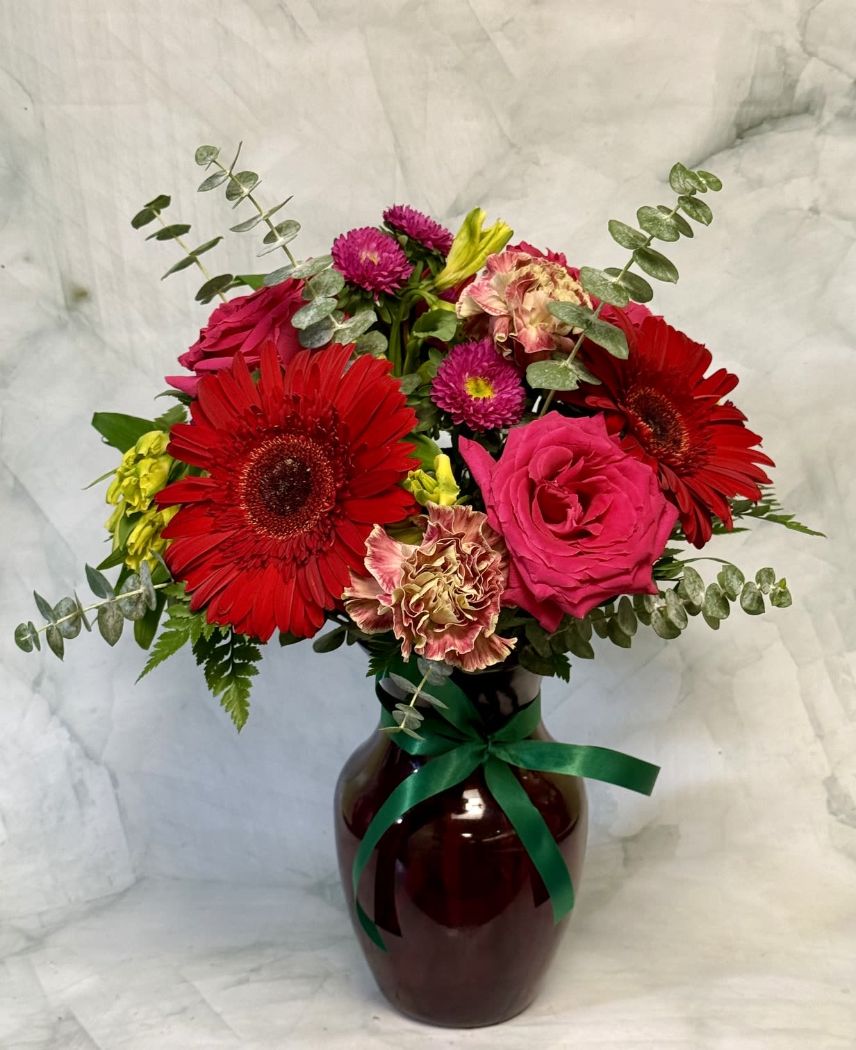 The Watermelon - Bright, high-contrast blooms make this flower arrangement an exuberant pick for a graduation celebration, a cheerful get-well gift, or a sunny summer surprise. The mix of red gerbera daisies and hot pink roses reads both joyful and slightly romantic, so the person you're thinking of will feel celebrated and seen-perfect for saying "congrats," "thank you," or "thinking of you" without being formal.  This bouquet combines vivid red gerbera daisies, bright pink roses, yellow alstroemeria, ruffled carnations in blush and cream, magenta button-style asters, and touches of green foliage for texture and lift. The flowers are gathered in a clear glass vase and finished with a green ribbon for a polished presentation; some versions include airy eucalyptus stems that give the arrangement a relaxed, hand-tied silhouette. Colors run from saturated reds and pinks to sunny yellow and crisp white accents for a lively, versatile palette.  Ideal for summer parties, Best Friends Day, Fourth of July gatherings, or a bedside pick-me-up, this arrangement arrives ready to display. Vase included; trim stems and freshen water every 2-3 days to extend enjoyment.
