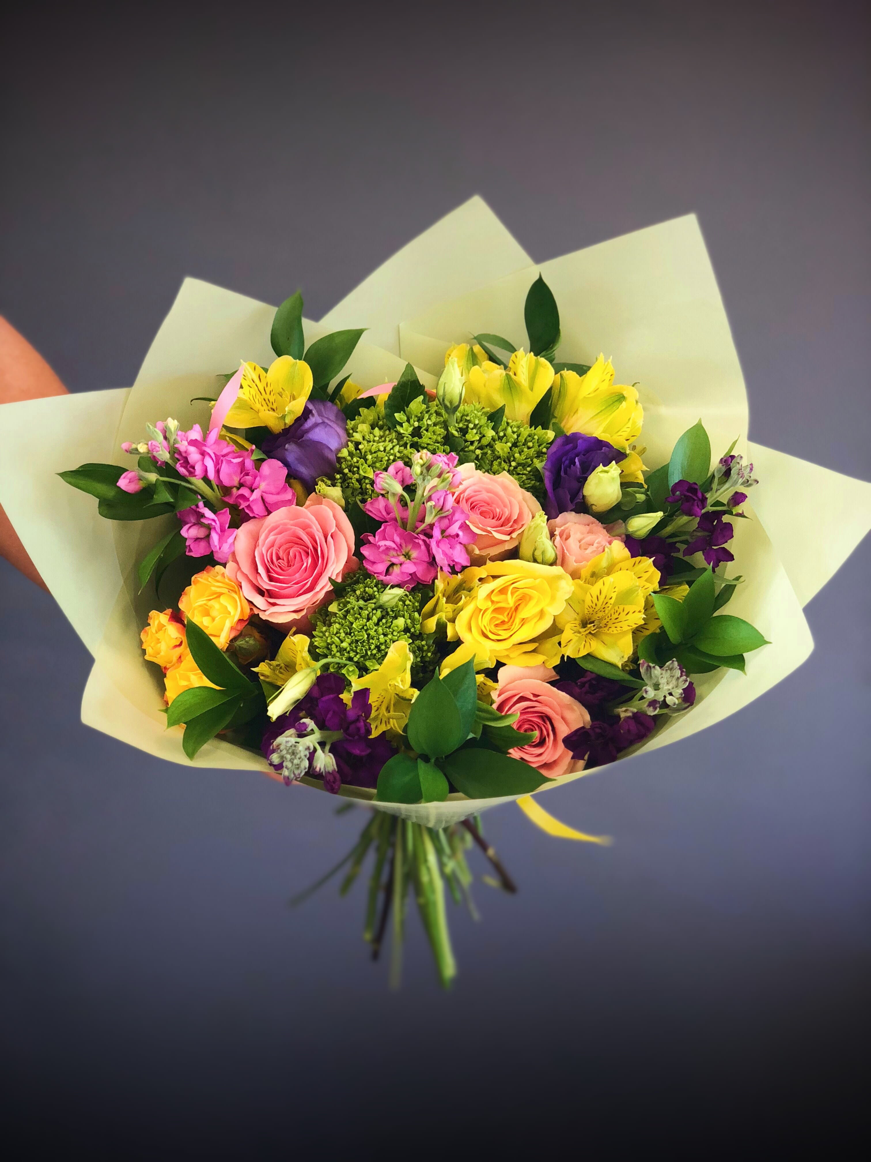 Mixed bouquet of pink, yellow, purple, and green flowers wrapped in pale paper