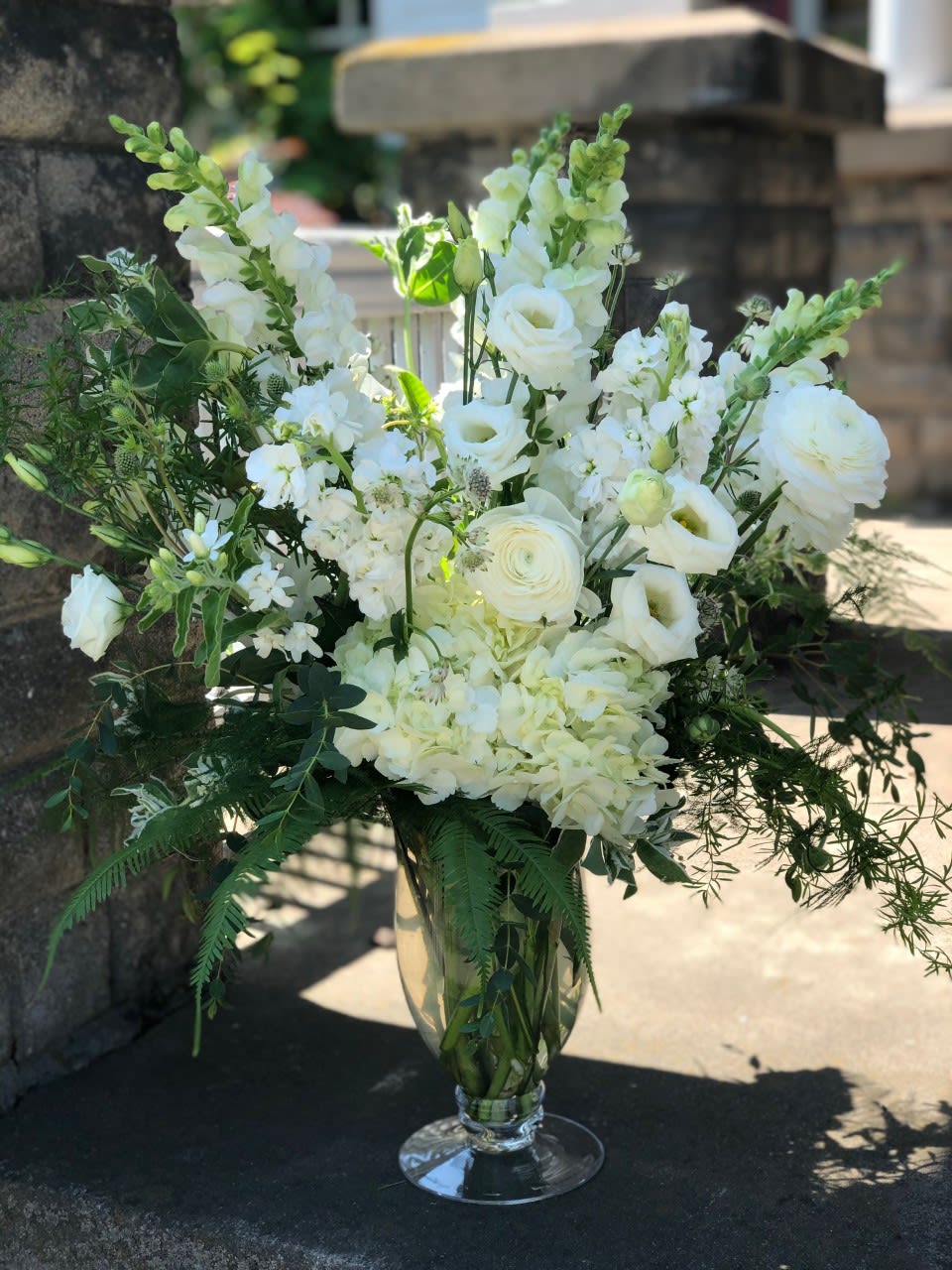 Tall all-white floral arrangement with roses, hydrangeas, and snapdragons in a clear glass pedestal vase