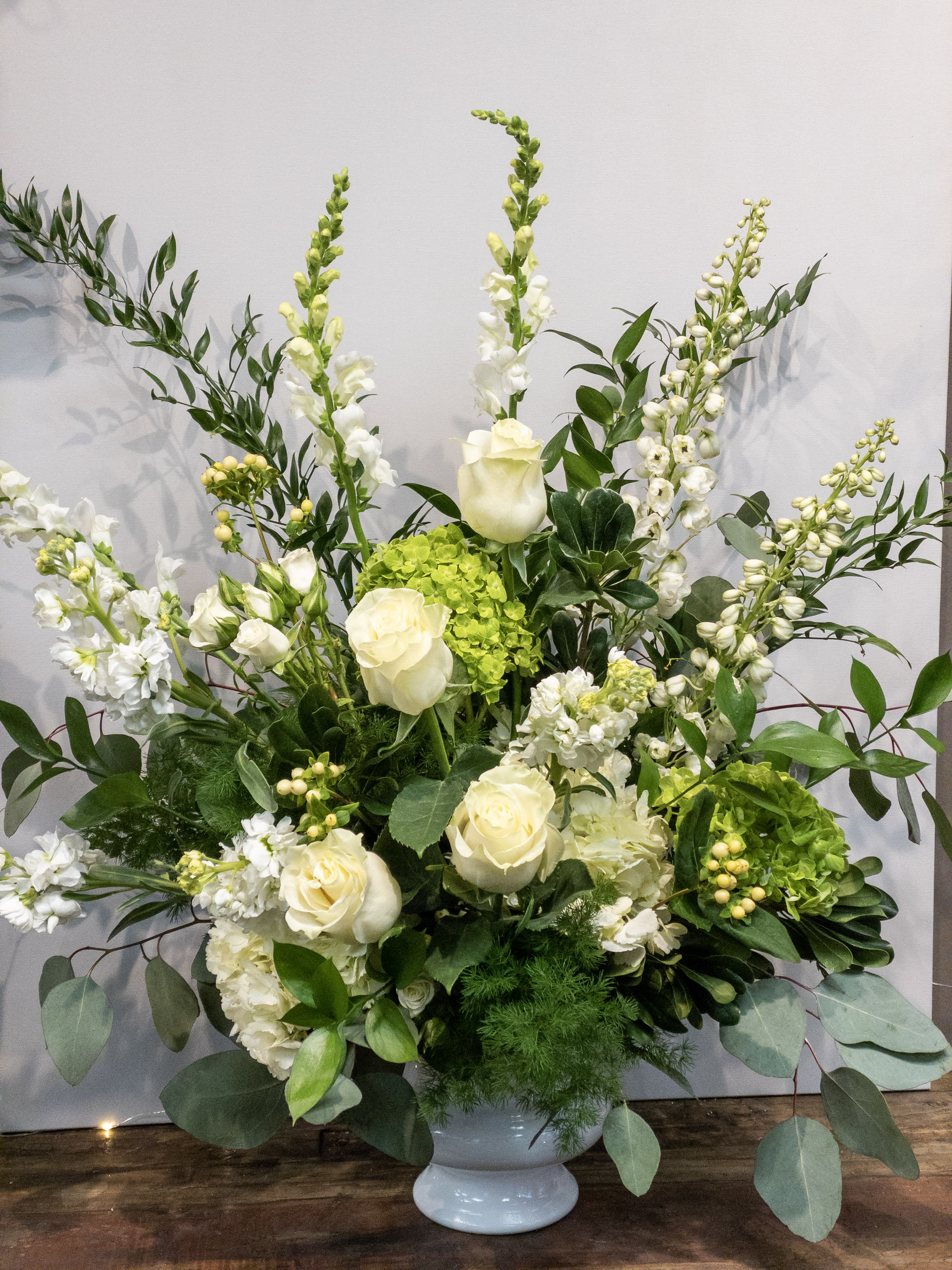 Tall white and green floral arrangement with roses, snapdragons, and hydrangeas in a white pedestal vase