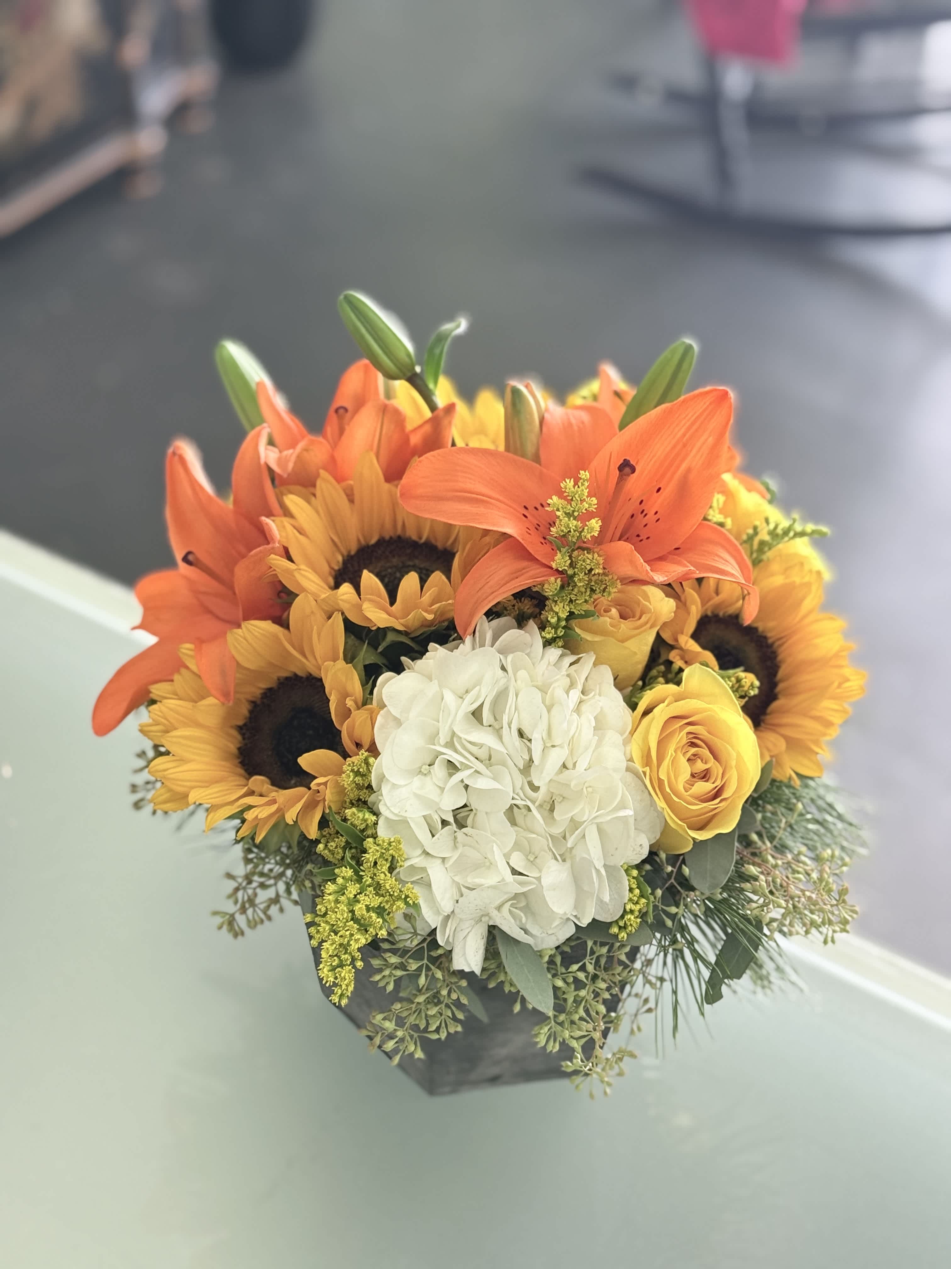 Orange lilies, sunflowers, a white hydrangea, and yellow roses in a low black container.