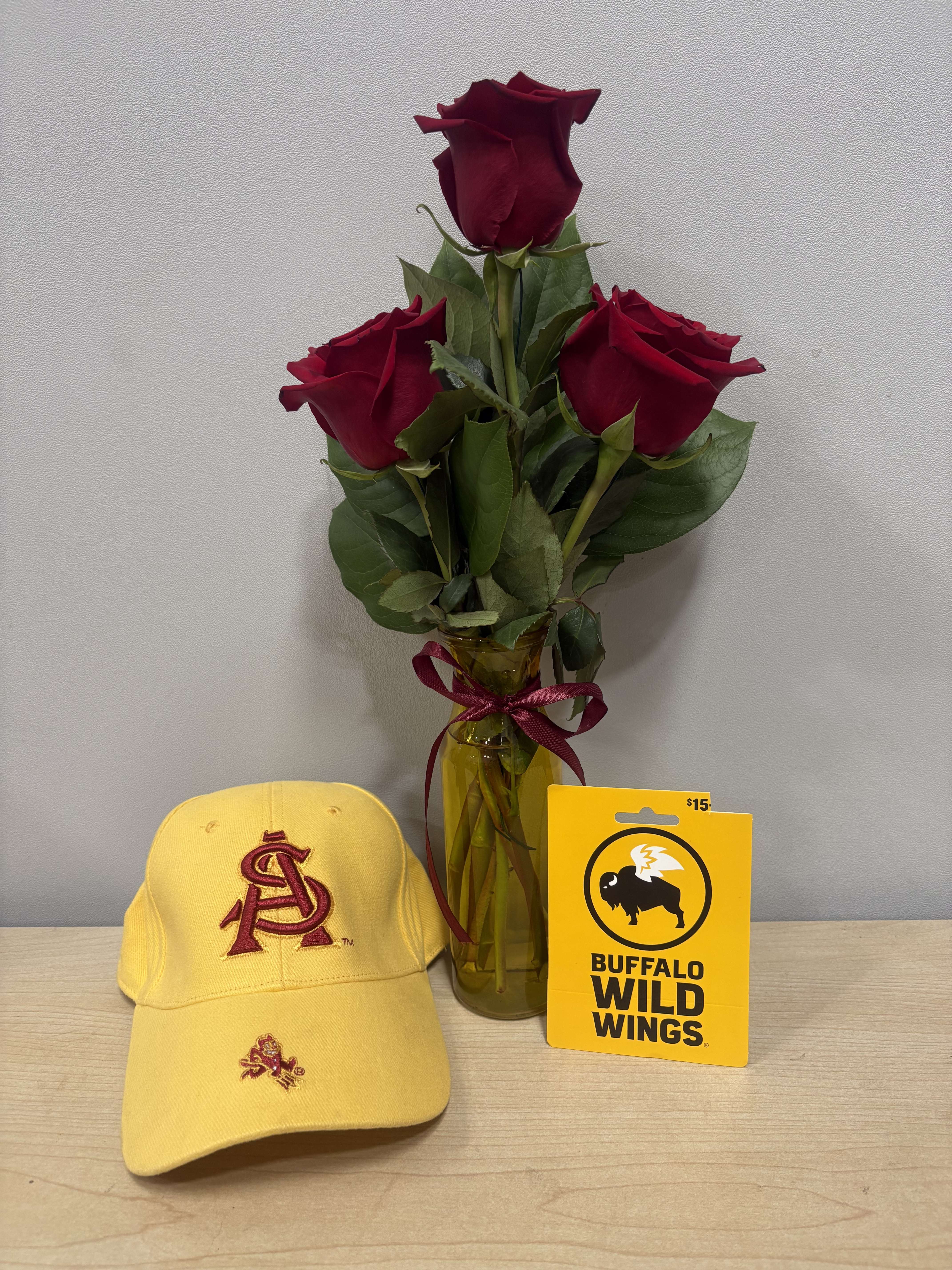 Three red roses in a glass vase with a ribbon, beside a yellow cap and gift card.