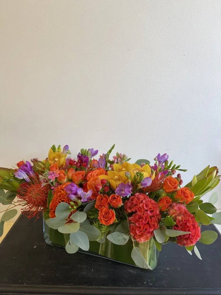 Low rectangular arrangement of orange, yellow, and purple flowers in a glass vase