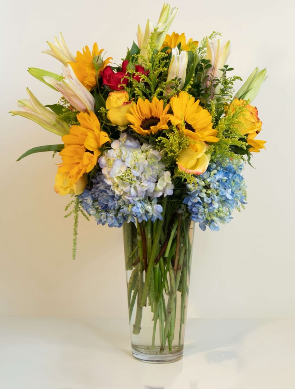Mixed bouquet in a clear glass vase with sunflowers, roses, lilies, and hydrangeas