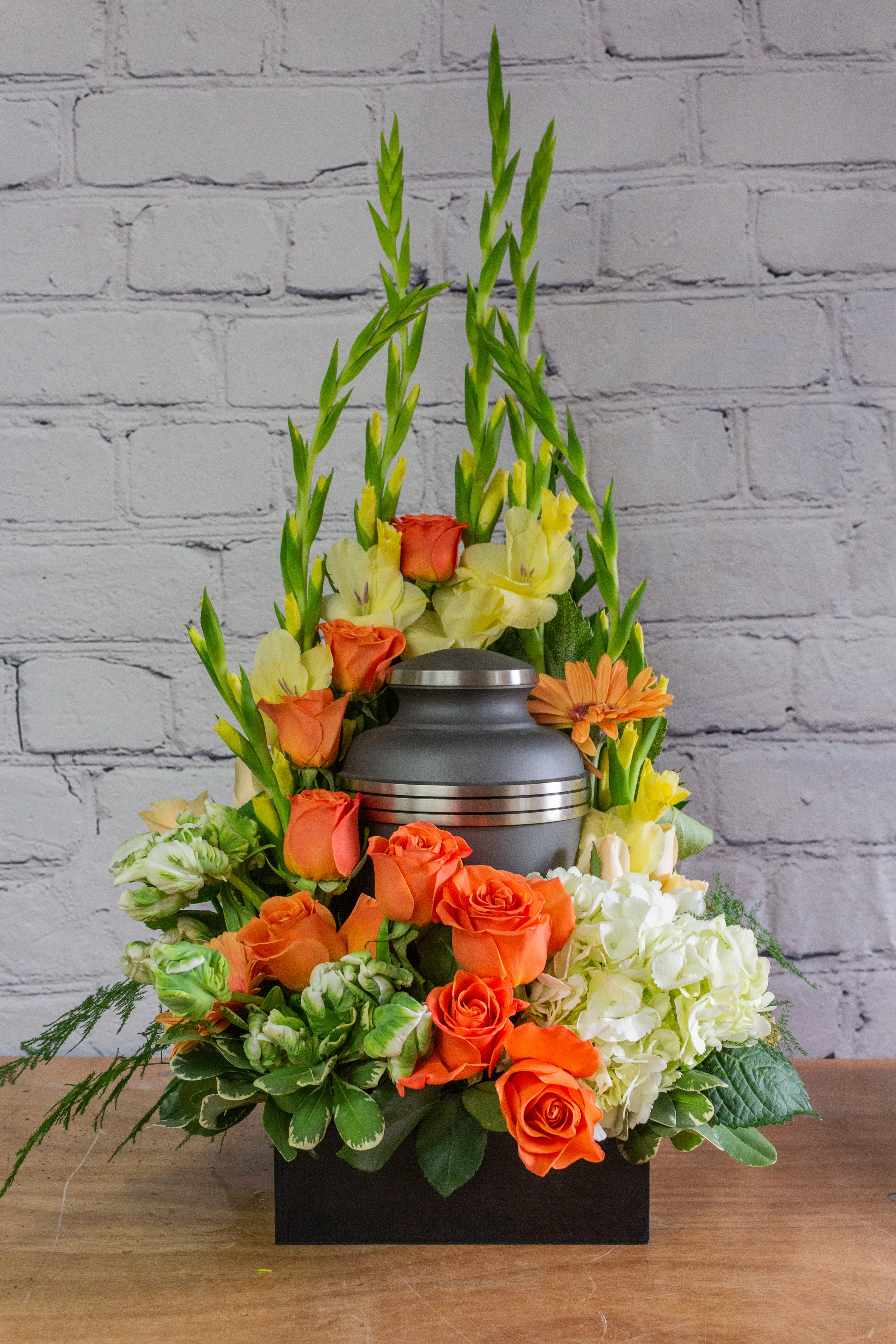 Orange roses and pale yellow flowers arranged around a gray urn