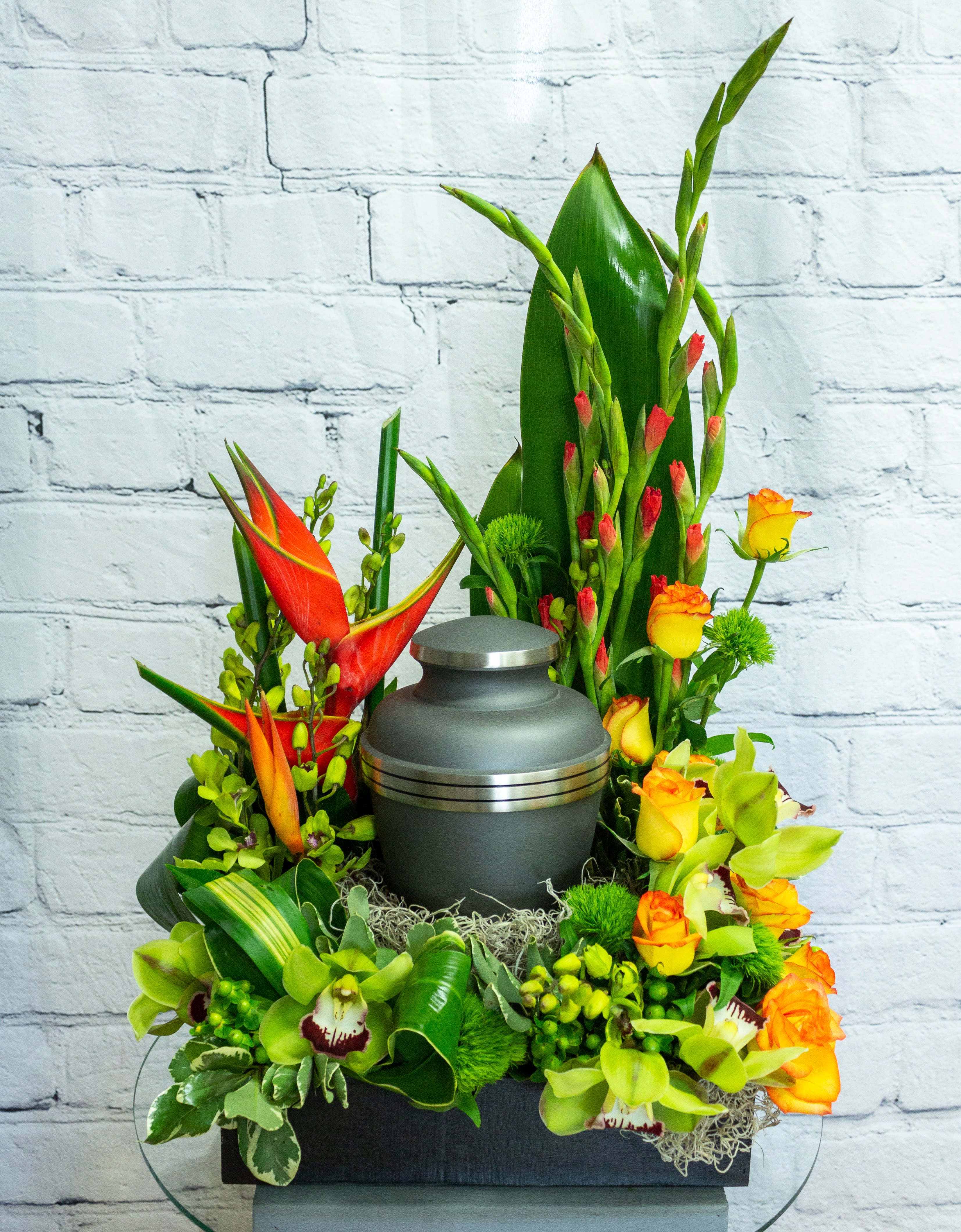Cremation urn surrounded by orange roses, green orchids, and tropical flowers in a tall arrangement