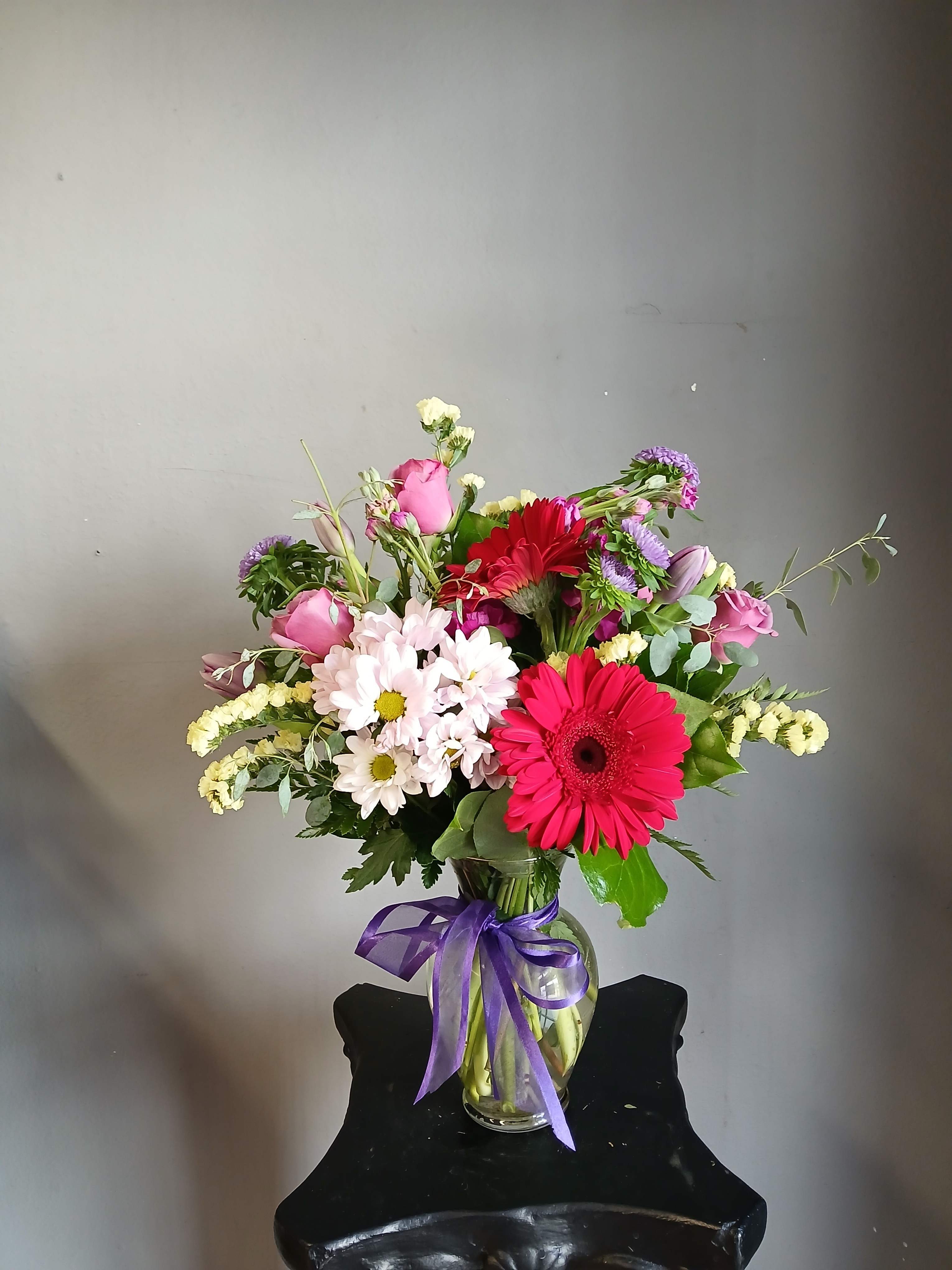 Mixed bouquet of pink, red, white, and purple flowers in a glass vase with a purple ribbon.