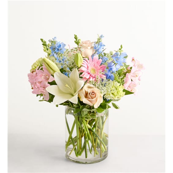 Mixed pastel bouquet in a clear glass vase with roses, lilies, and blue flowers