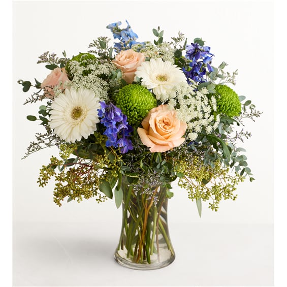 Mixed bouquet of peach roses, white gerberas, and blue flowers in a glass vase