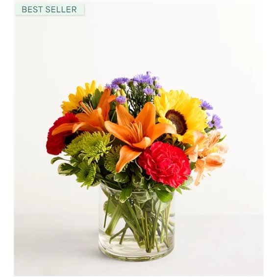Mixed bouquet of orange lilies, sunflowers, and red carnations in a glass vase