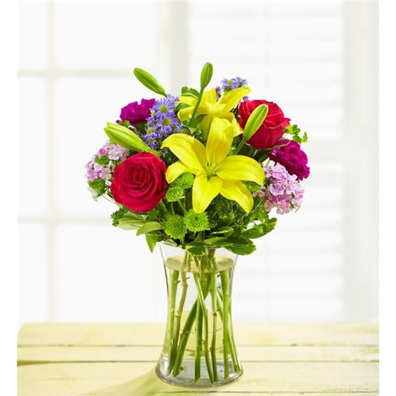 Bouquet of yellow lilies, red roses, and purple flowers in a clear glass vase