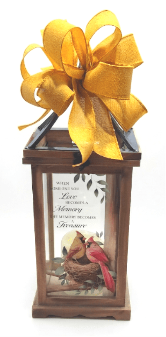 "Memory Becomes a Treasure" Lighted Lantern - A warm wooden lantern topped with a large yellow bow offers a quiet, comforting presence-perfect as a sympathy gift or remembrance keepsake to honor a loved one. The printed quote reads "When Someone You Love Becomes a Memory The Memory Becomes a Treasure" lighted lantern reads as a thoughtful memorial piece for funerals, anniversaries of passing, or for placing on a mantel where someone dear is remembered; the printed quote and bird artwork gently convey consolation and enduring memory.  The lantern features warm brown wood, clear glass panels, and decorative bird artwork showing two cardinals in a nest. A flameless LED candle sits within the wooden frame (built-in auto timer: 6 hours ON / 18 hours OFF) so the soft glow can be left safely on display; three AAA batteries are included.  Dimensions are W: 6.25", H: 16.5", D: 5.5", and it's intended for indoor display only.  This keepsake-style piece makes the person you're honoring feel remembered and seen-a quiet way to express sympathy or mark a meaningful date. Order with confidence for delivery to a home or gathering space where a lasting, elegant reminder of love is needed.