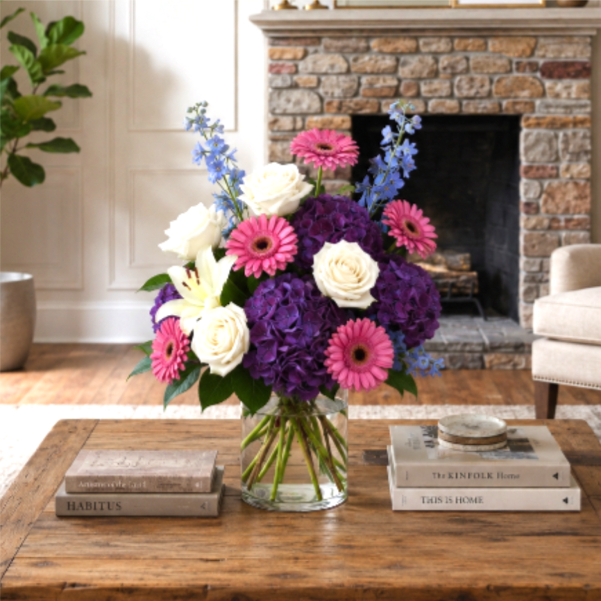 Bouquet of pink gerbera daisies, white roses, and purple hydrangeas in a glass vase