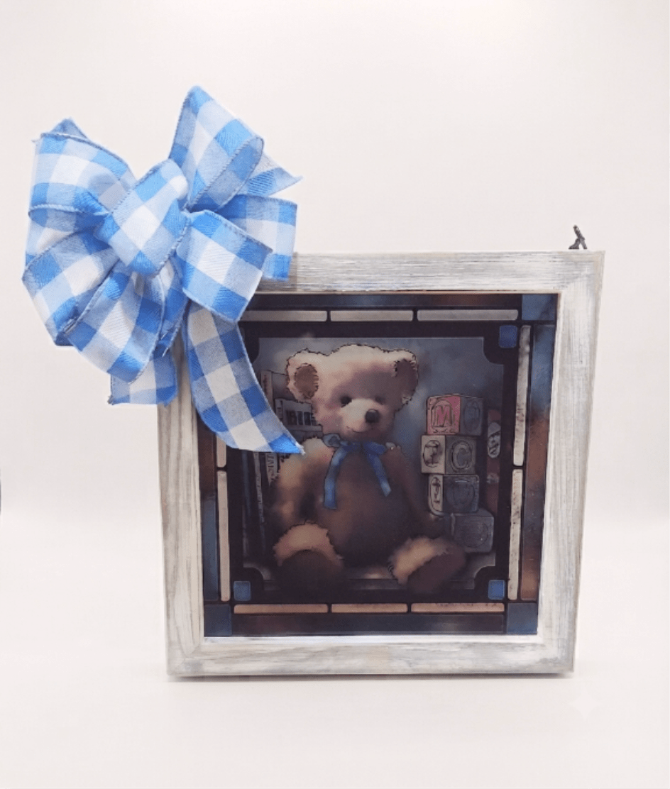 "ABC Bear for a Boy" Stained Glass - A sweet plush teddy bear with a blue ribbon is the heart of this nursery-ready stained glass panel - a charming keepsake flower arrangement alternative perfect for a new baby boy or a birthday gift. The square art hanging reads like a vintage nursery window: a tan bear seated before pastel alphabet blocks and stacked children's books framed by geometric panes in deep blue, burgundy, and neutral tones that give the piece depth and a warm, comforting glow.  Set in a distressed white wood frame and suspended by a simple metal chain, the piece is finished with a matching bow and presented ready to hang or display on a tabletop easel. (Tabletop Easel provided with delivery or pickup.) The printed glass uses fade-resistant inks for lasting color, and the metal chain and wood frame create a sturdy, decorative accent that looks especially lovely back-lit by natural light.  A thoughtful way to welcome a newborn or celebrate a little one's birthday, he will feel loved and seen every day. Includes ready-to-hang hardware and a tabletop easel is included.  10 x 10 