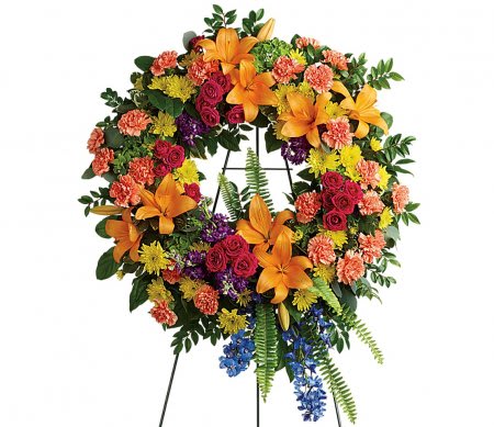 Bright multicolor standing wreath with lilies, roses, carnations, and mums on a metal easel.