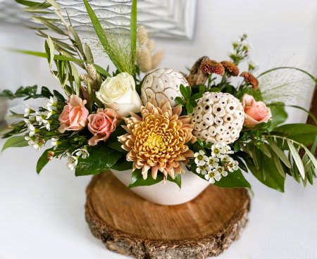 Low arrangement of peach roses, white rose, yellow chrysanthemum and white pods in a bowl vase