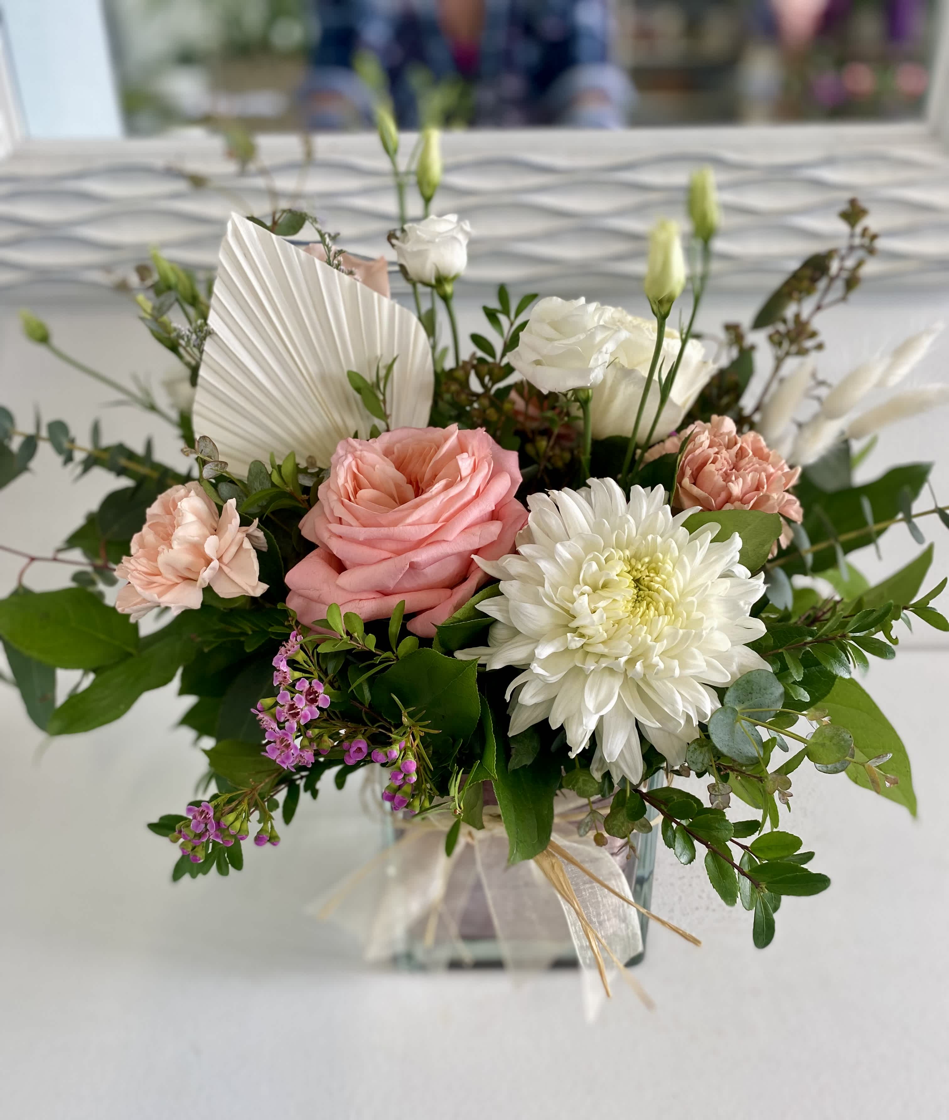 Pink roses, peach carnations, and white blooms in a glass cube vase with ribbon.