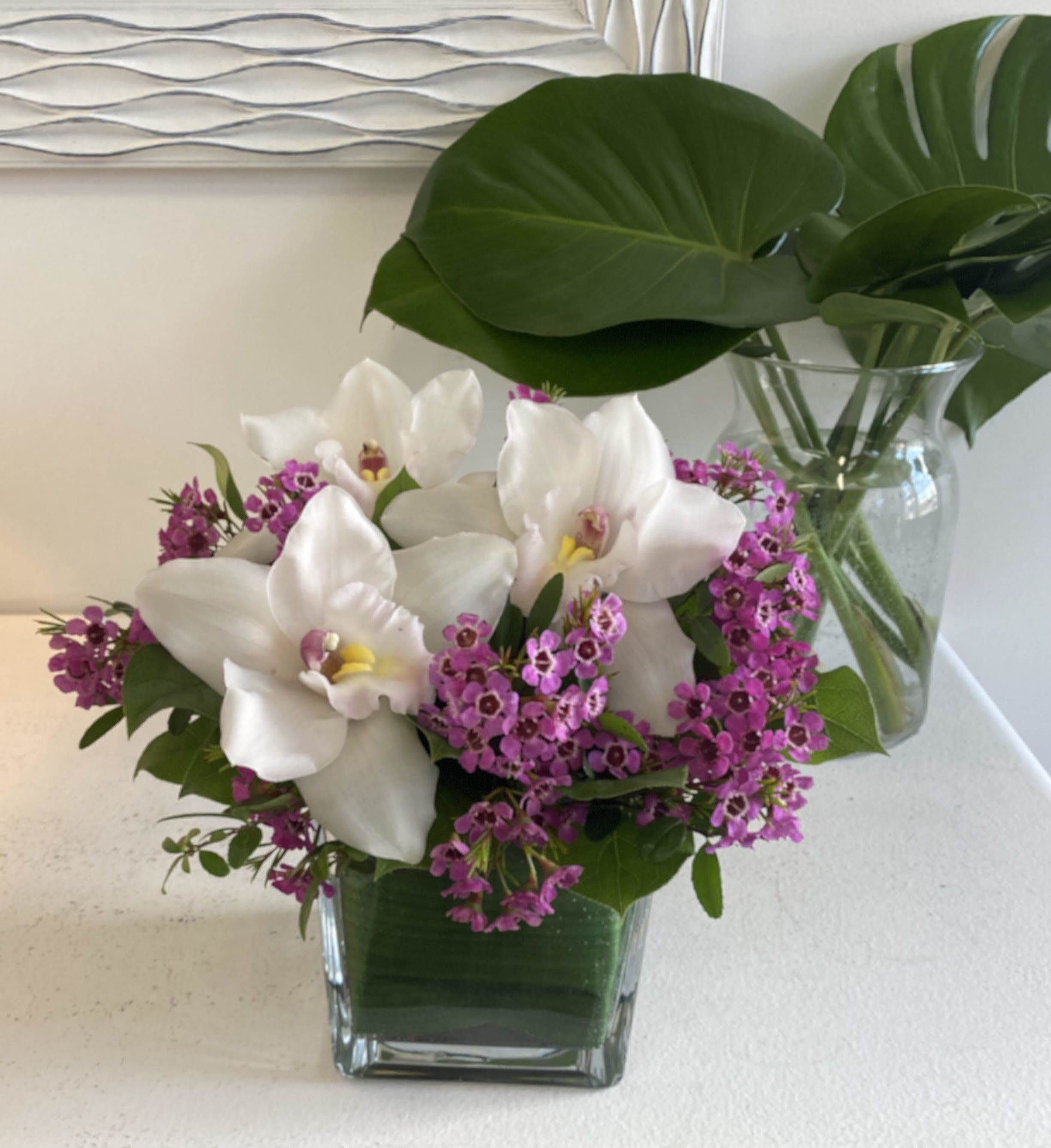Compact arrangement of white orchids and small purple flowers in a square glass vase