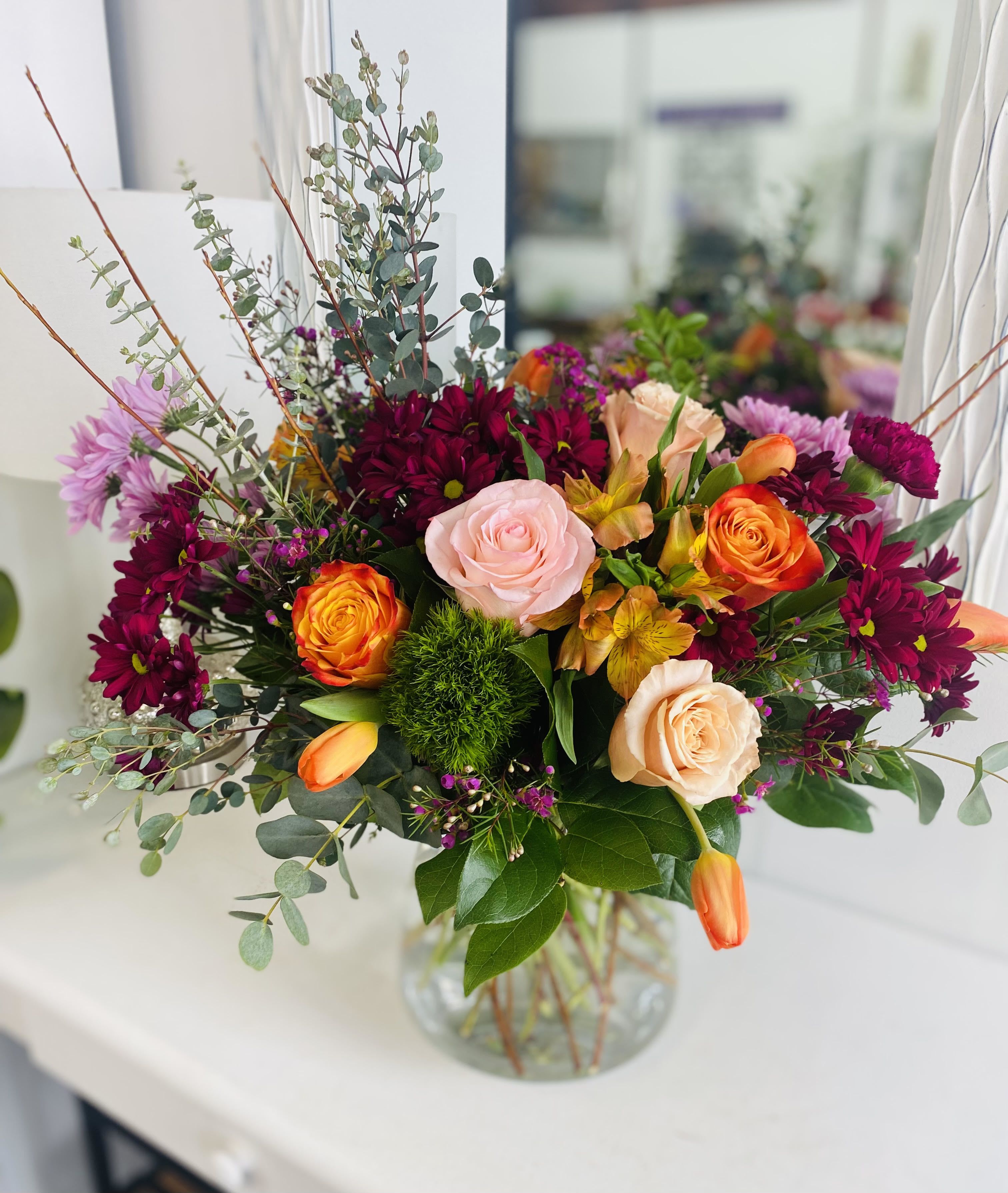 Vibrant arrangement of pink and peach roses, orange tulips, and burgundy mums in a clear glass vase