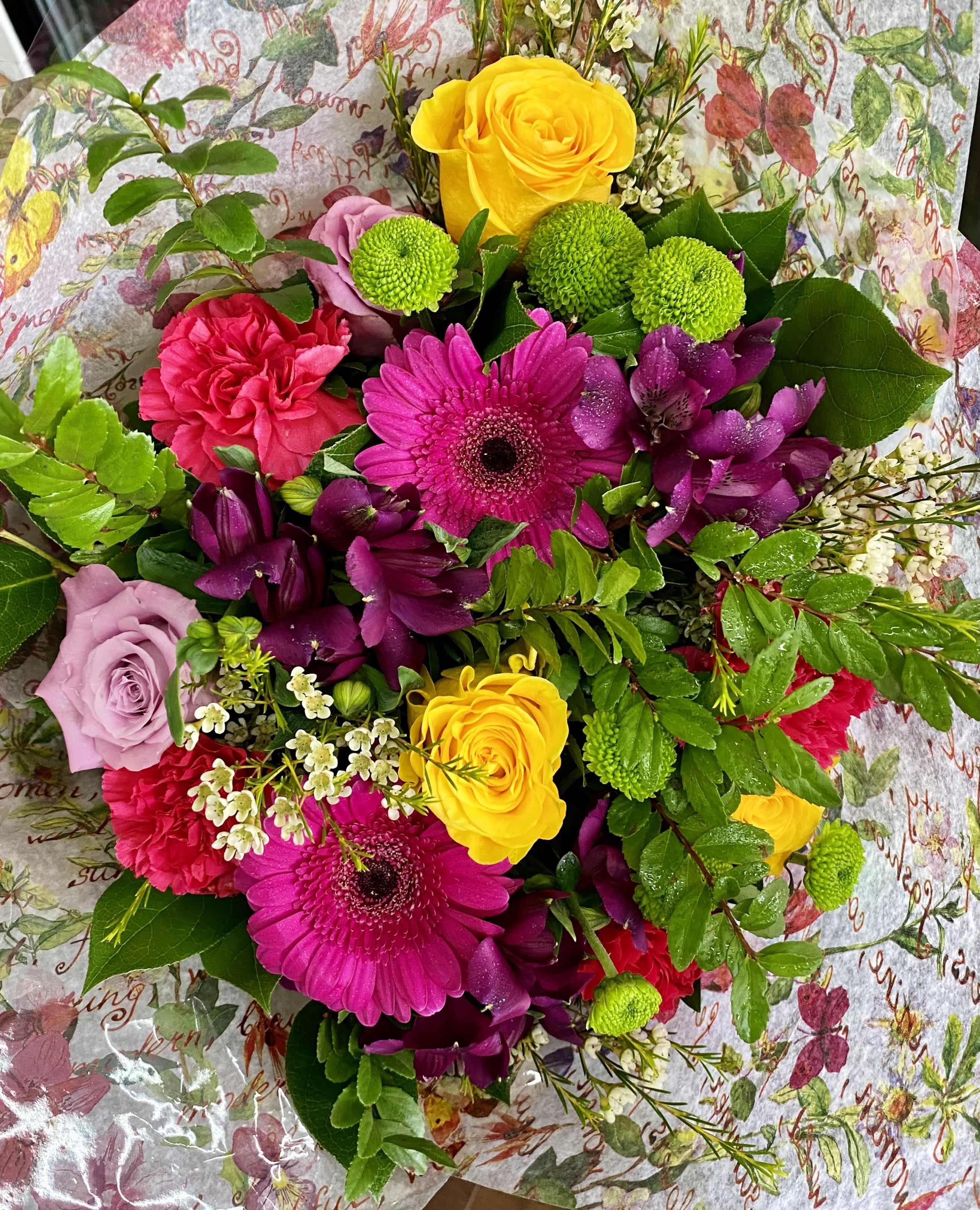 Bright hand-tied bouquet with pink gerbera daisies, yellow roses, green mums, and purple flowers in floral wrap