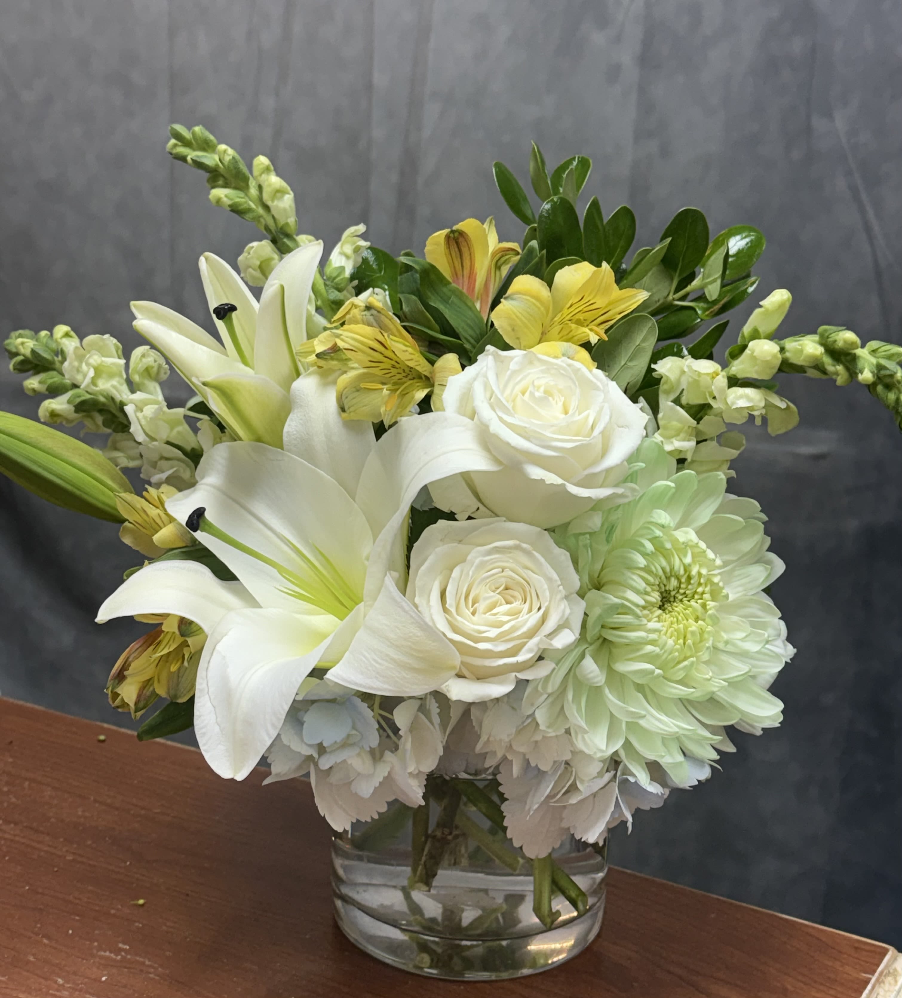 Serenity Bloom - A graceful arrangement blending soft blue hydrangeas, elegant white lilies, creamy white roses, sage green mums, and delicate white carnations. This serene design evokes calm, purity, and natural beauty — perfect for thoughtful gifts, sympathy gestures, or peaceful home décor.