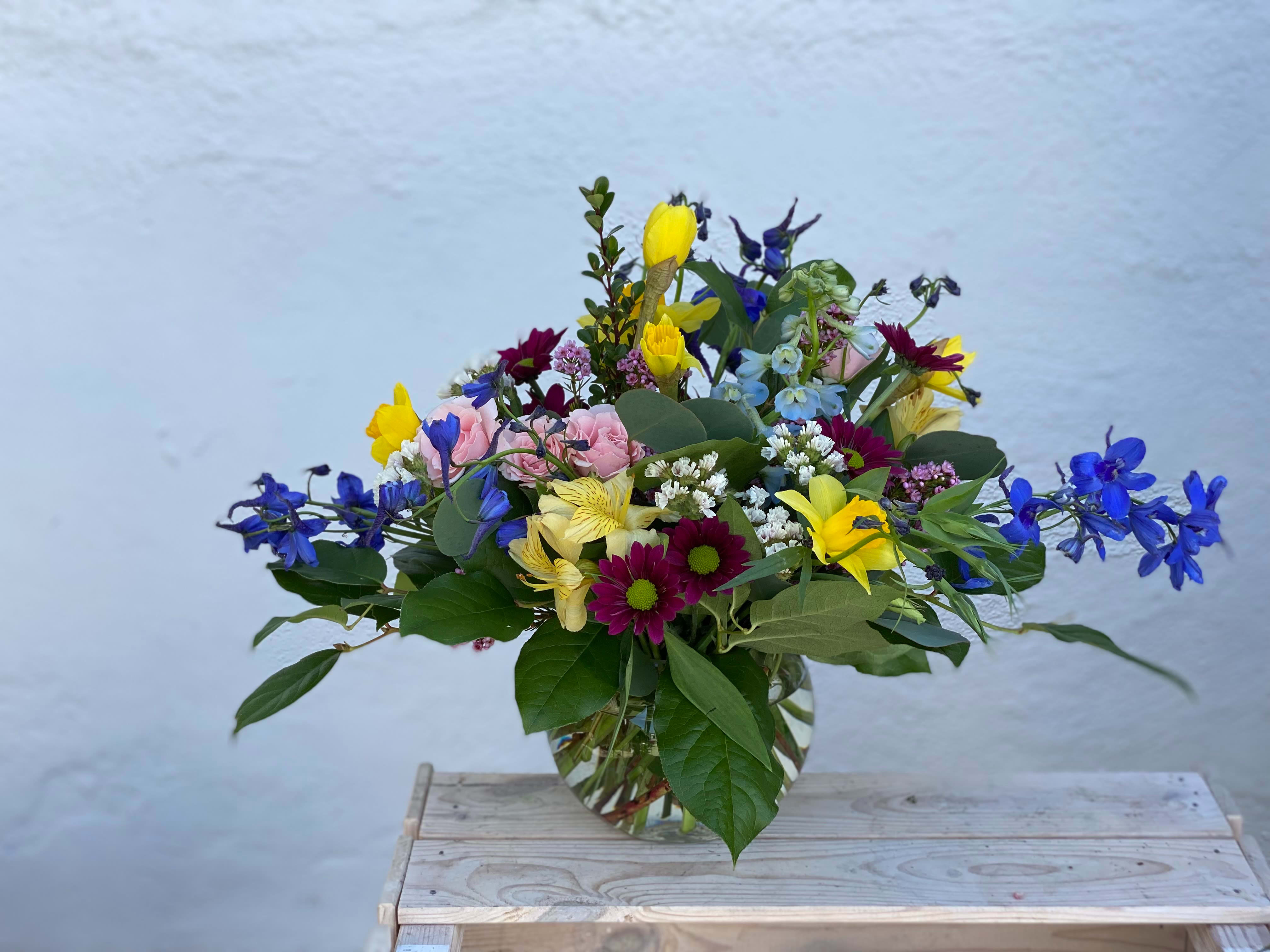 Mixed bouquet in a clear glass vase with blue, yellow, pink, and purple flowers.