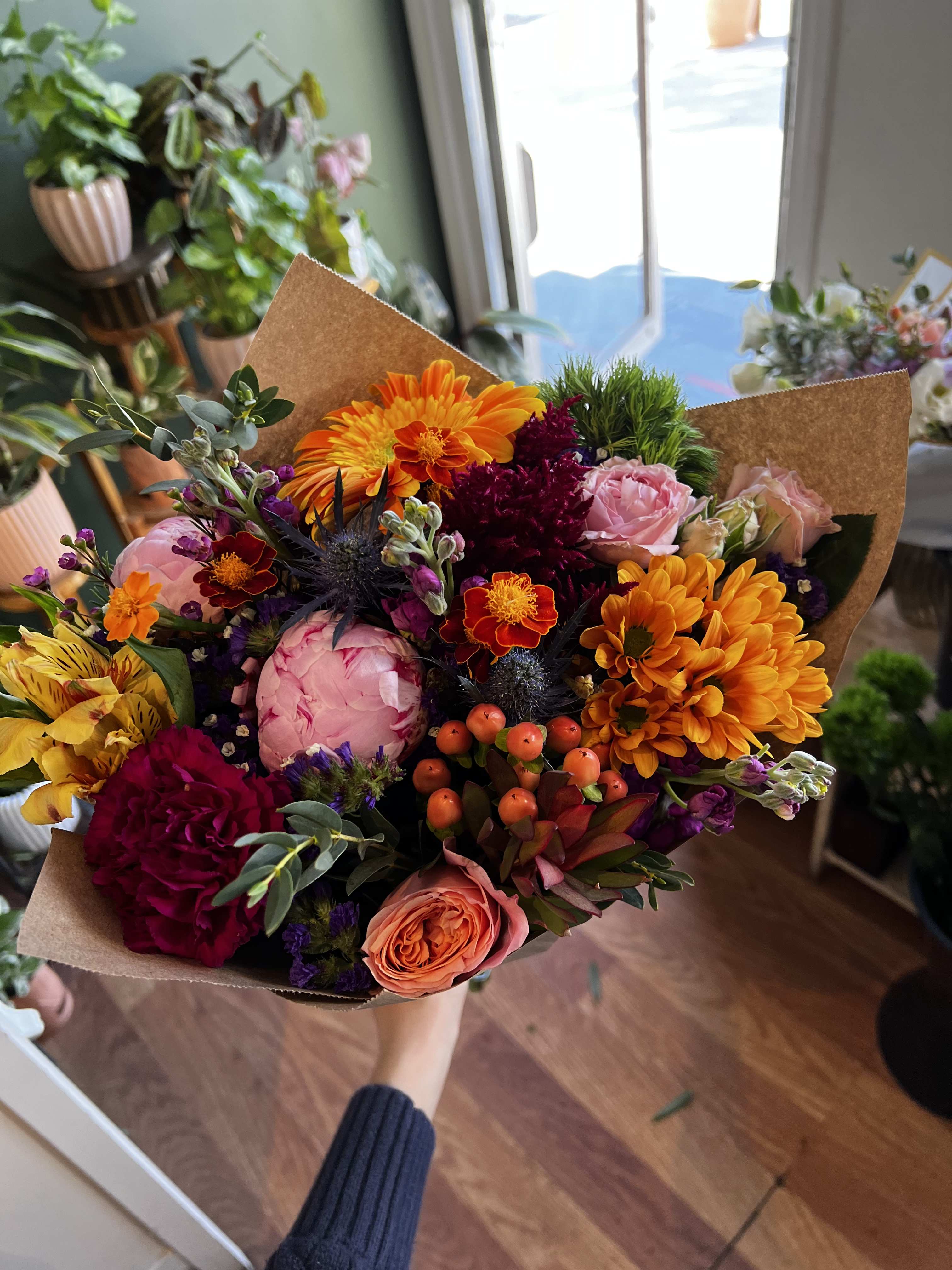 Handheld bouquet of mixed orange, pink, and burgundy flowers in brown paper
