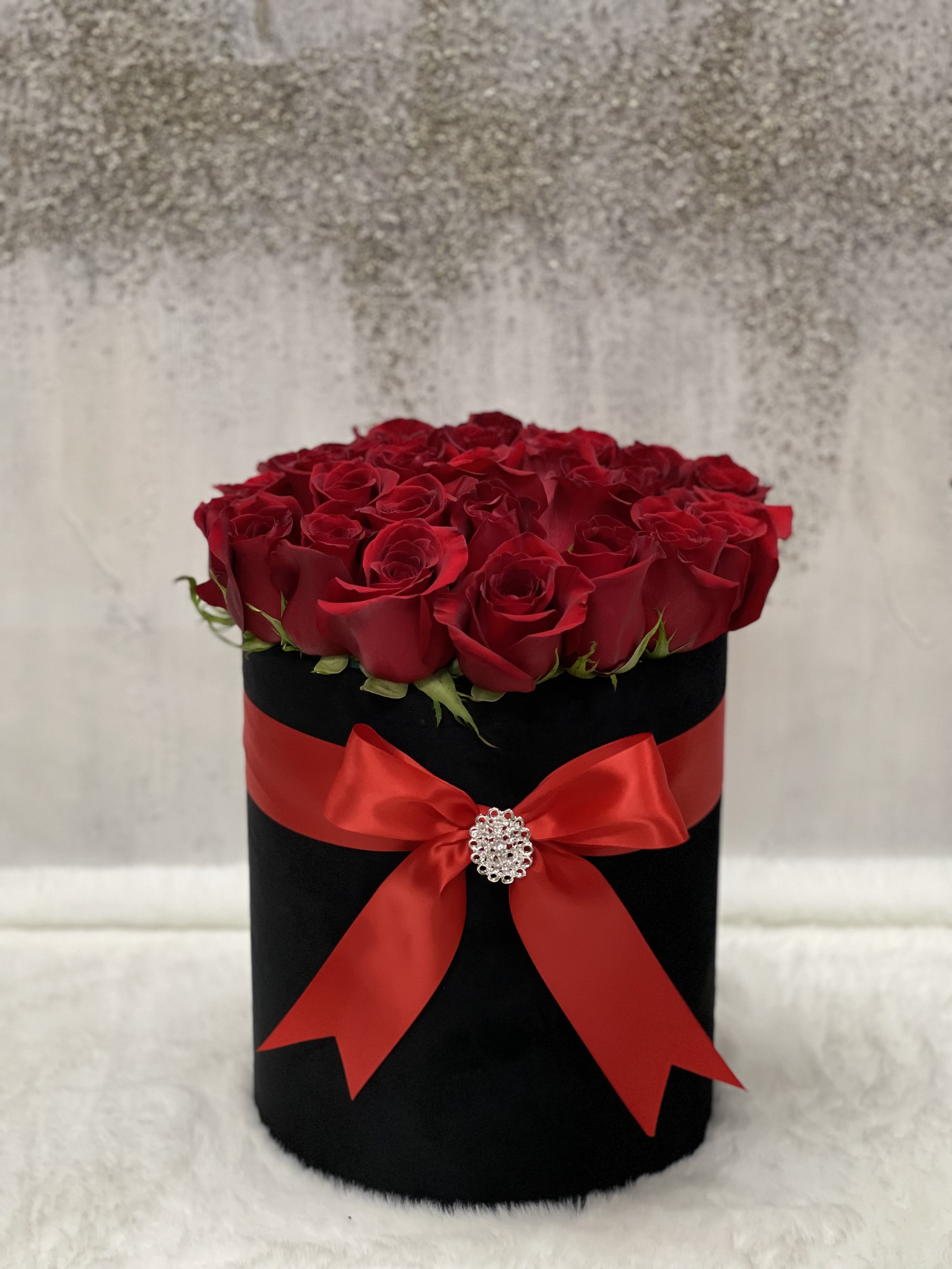 Round black hatbox of red roses wrapped with a red satin bow and jeweled brooch.